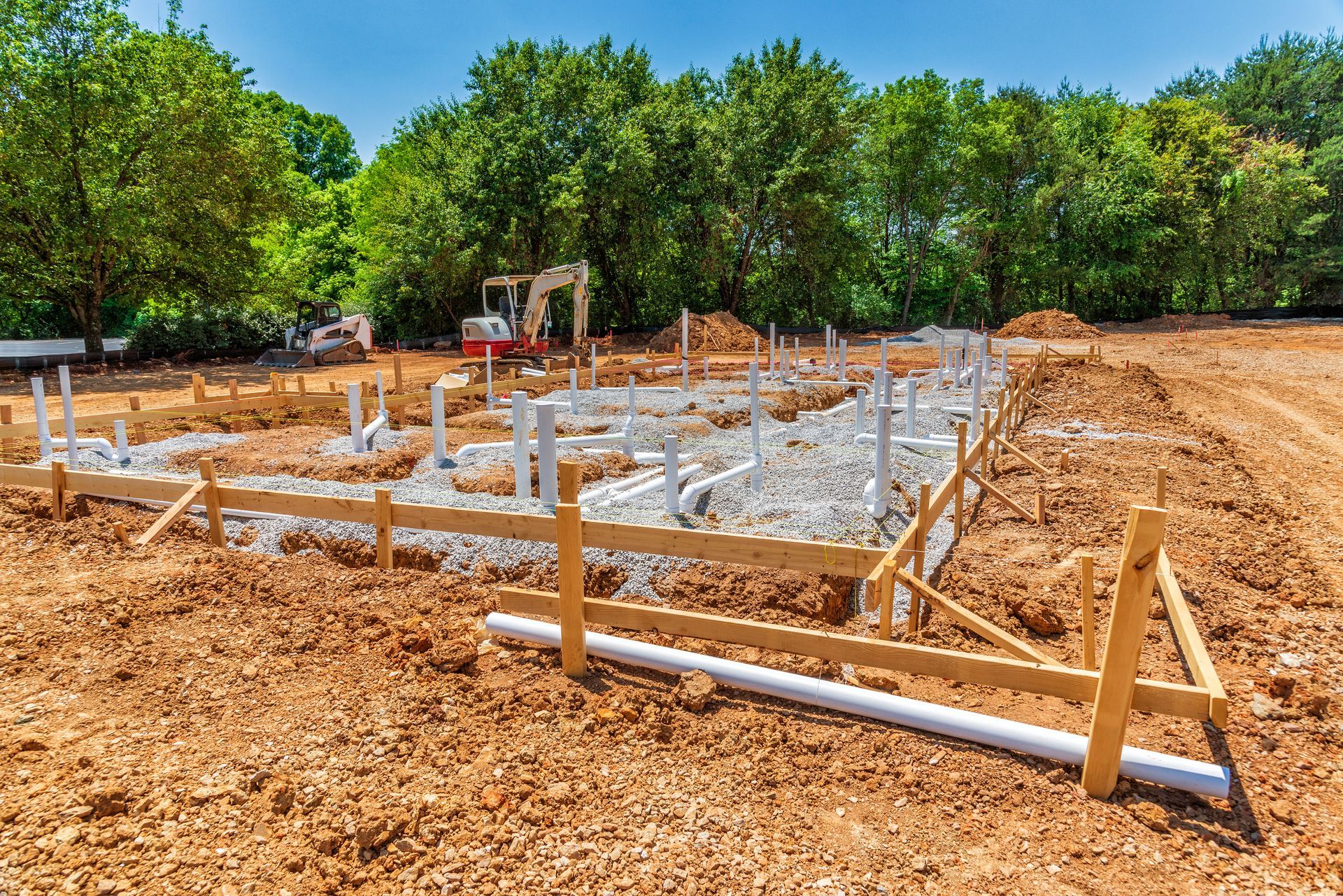 Construction site with wooden forms and plumbing pipes laid out in dirt. Trees in the background.