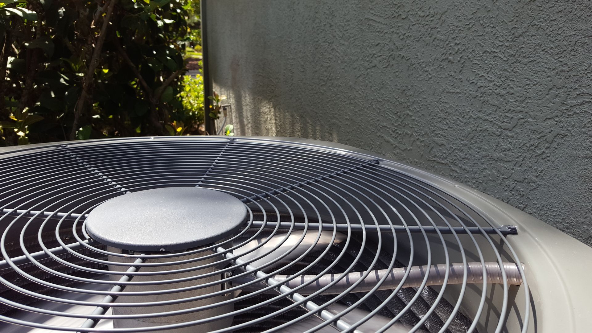 Air conditioner unit against a textured stucco wall; metal fan grill.