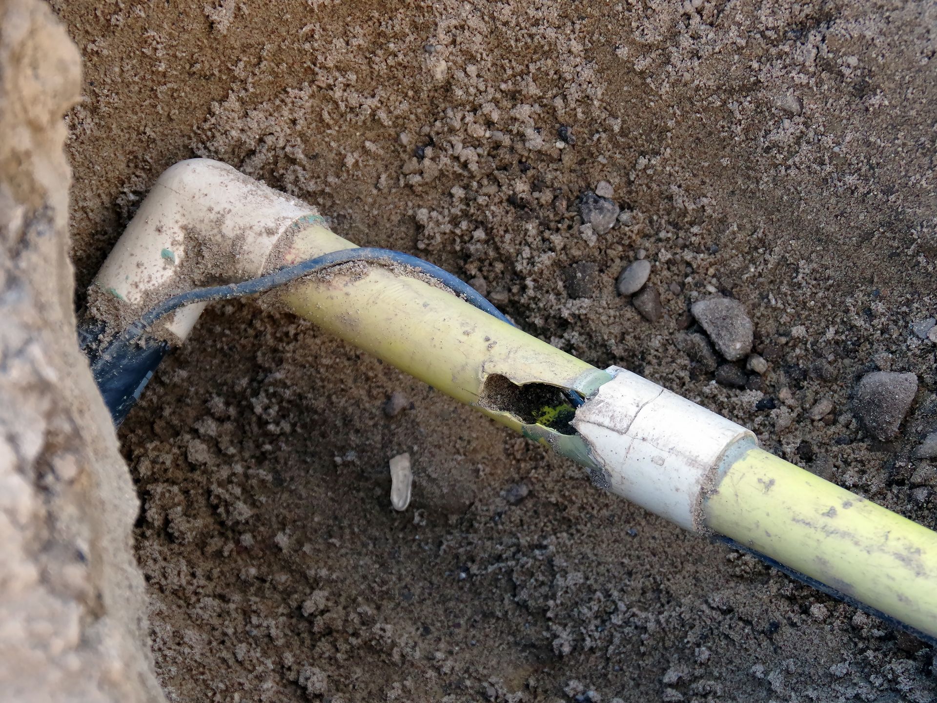 Broken yellow plastic pipe in dirt, with hole and exposed wires.