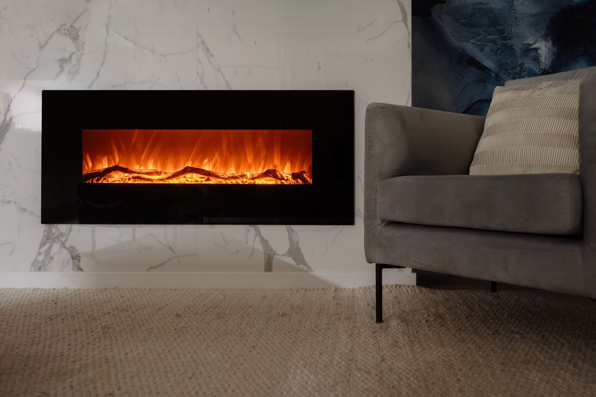Electric fireplace with orange flames against a marble wall, beside a gray armchair.