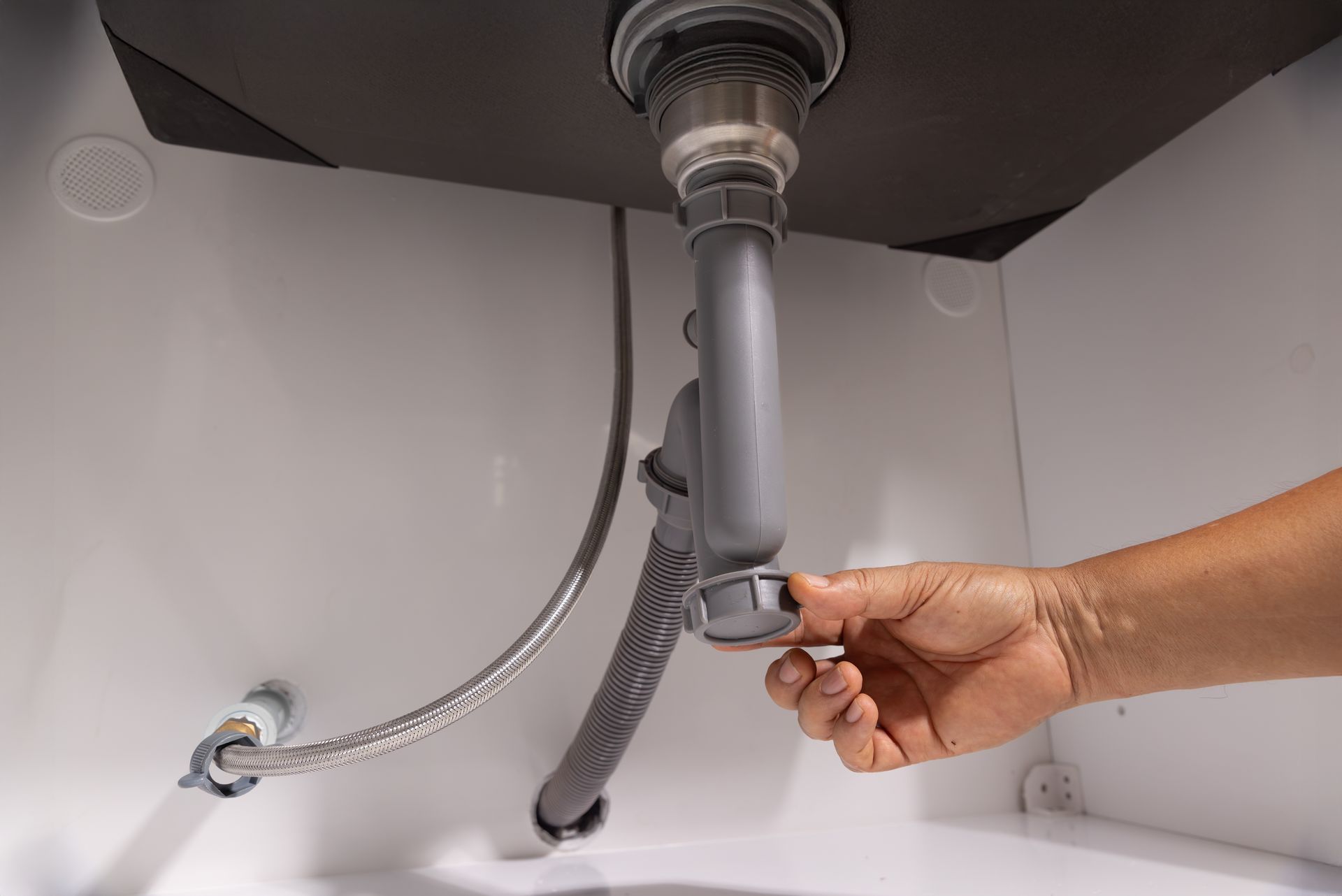 Hand unscrewing a P-trap under a white sink. Gray plumbing and a silver strainer are visible.