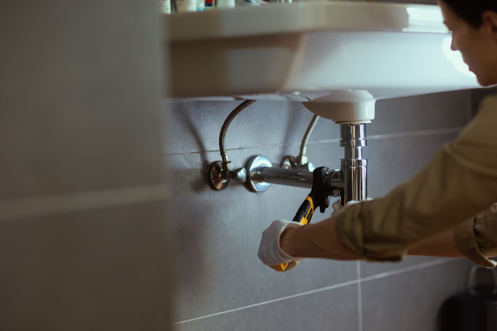 Plumber using a wrench under a white sink, working on plumbing. Gray tile background.