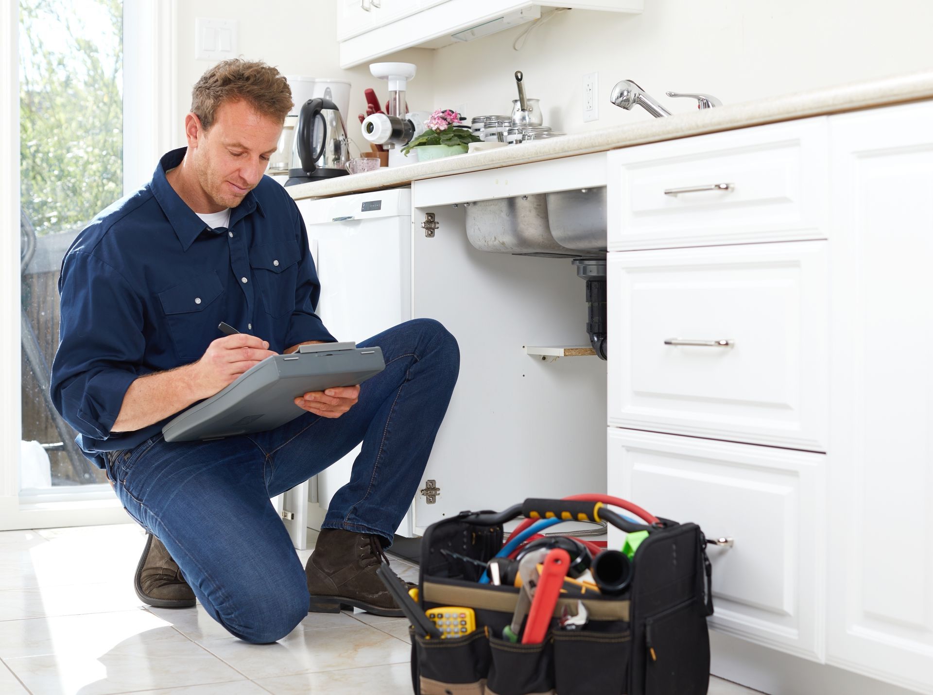 Plumber kneeling under kitchen sink, inspecting pipes and writing on a clipboard. Toolbox in foreground.