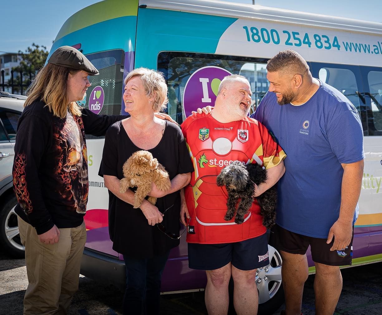 Four people stand outside by a colorful van, smiling as two of them hold small dogs.
