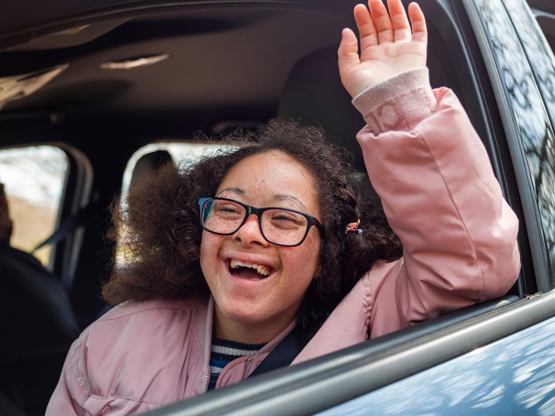 Person waving from car window, smiling, wearing glasses and pink jacket.