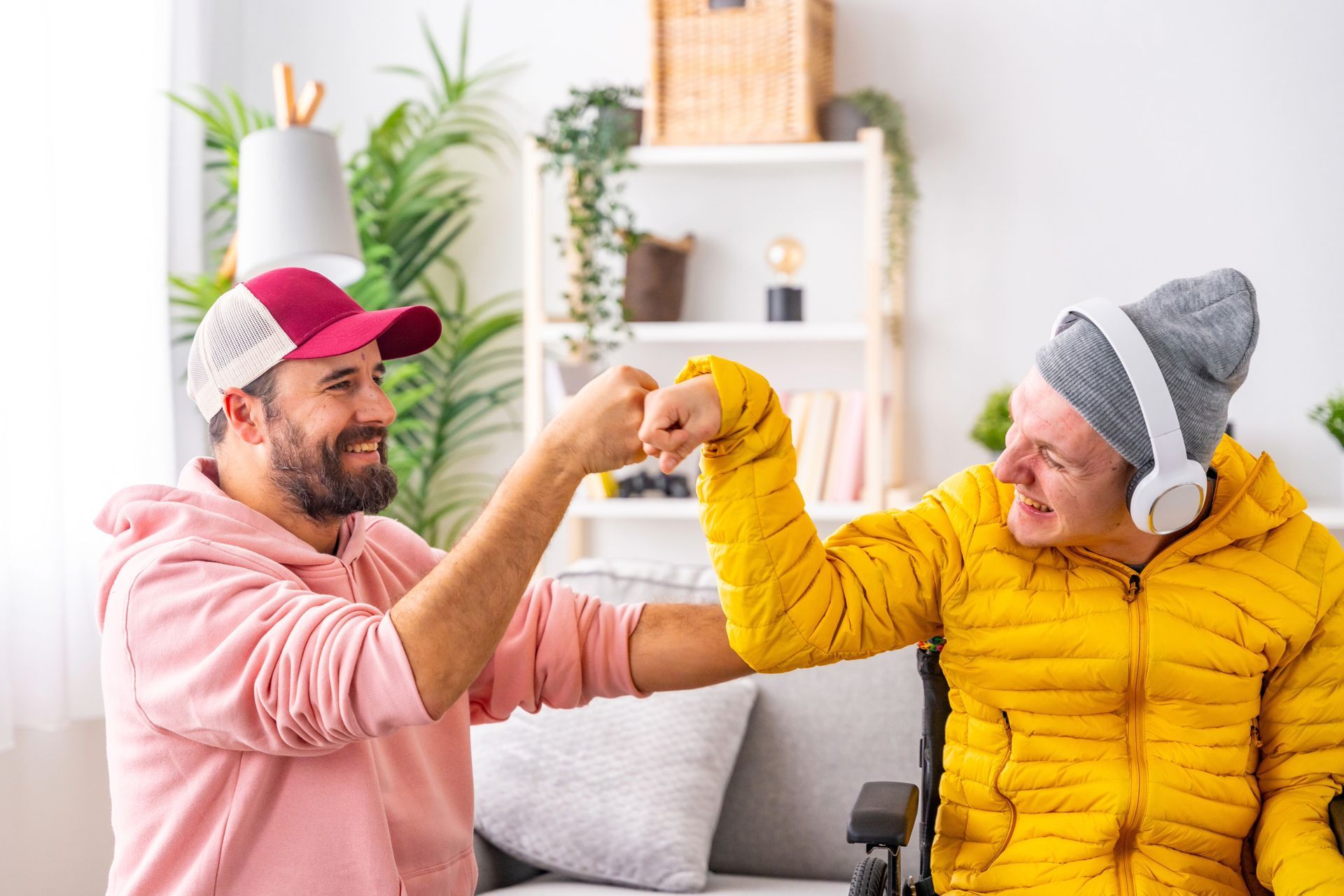Two men bump fists, one in a wheelchair, indoors. One wears a pink hoodie and hat, the other a yellow jacket and headphones.