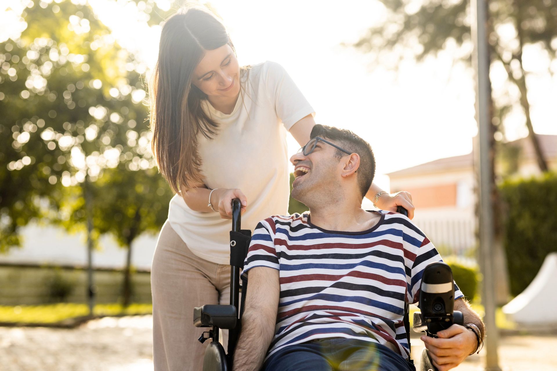 Woman smiling, pushing a wheelchair with a man who is also smiling, outside in the sunshine.
