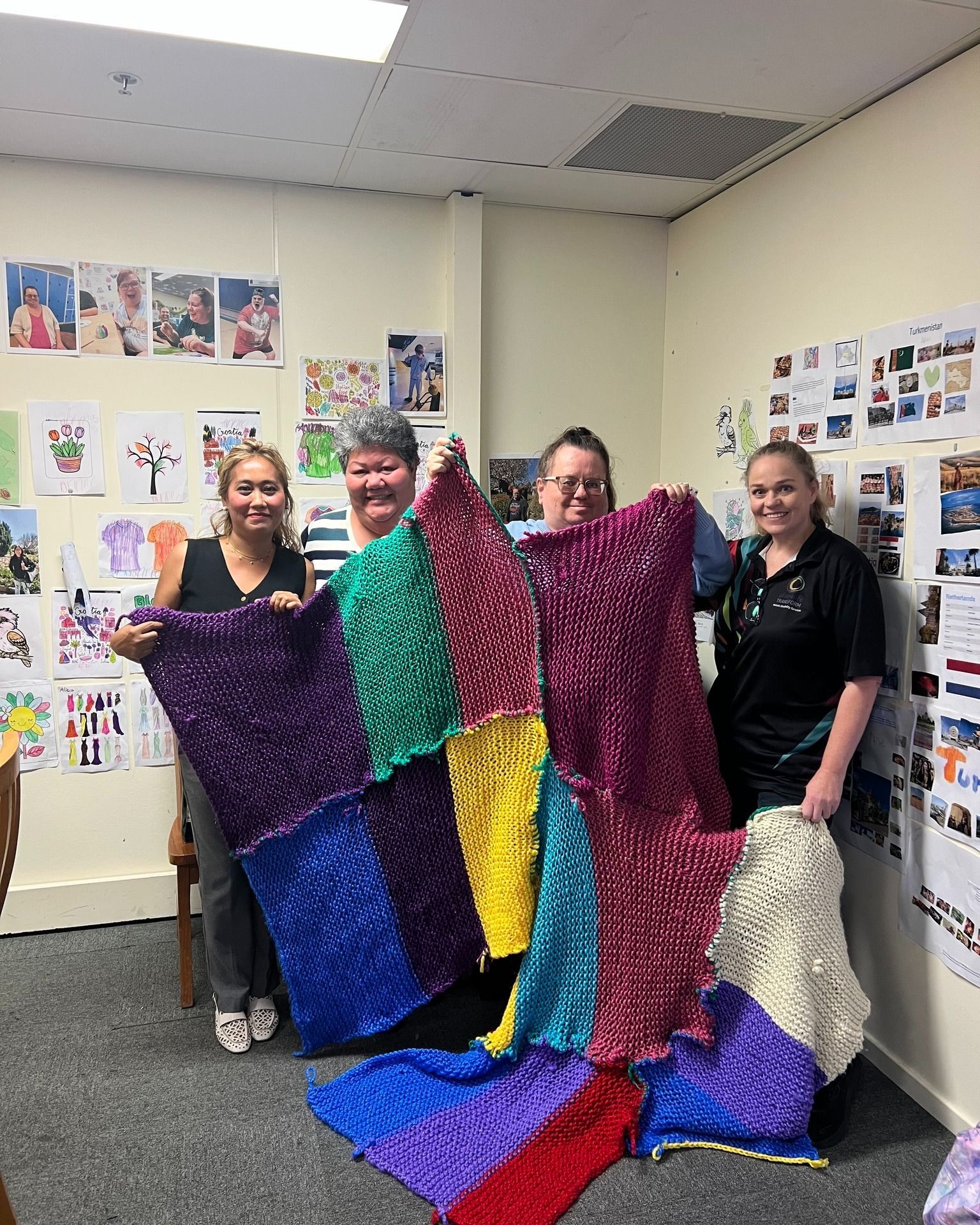Four people holding up a large patchwork blanket, smiling, in a room with artwork on the walls.