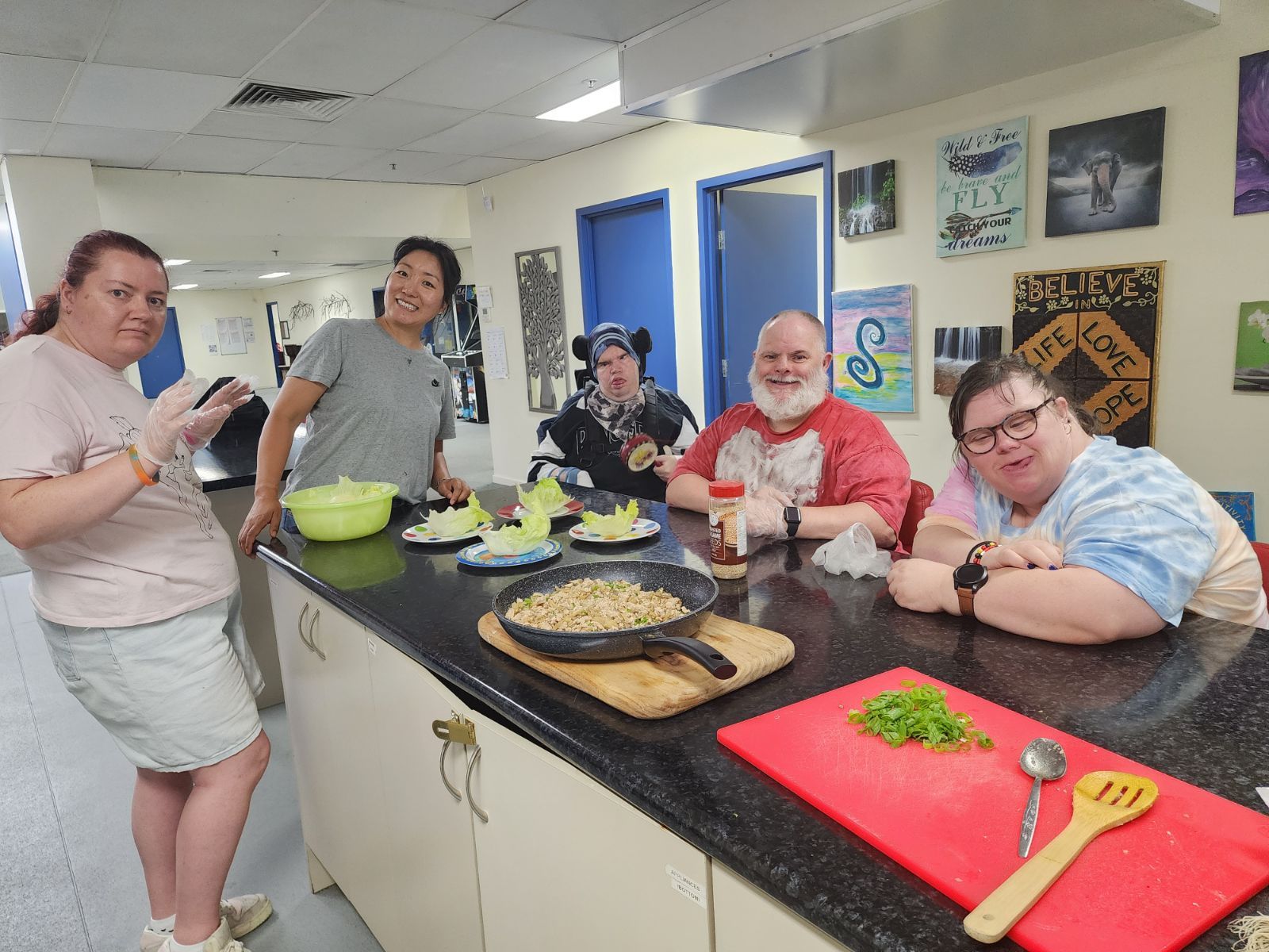People cooking together at a counter. Lettuce, a frying pan, and chopped vegetables are visible. Joyful expressions.