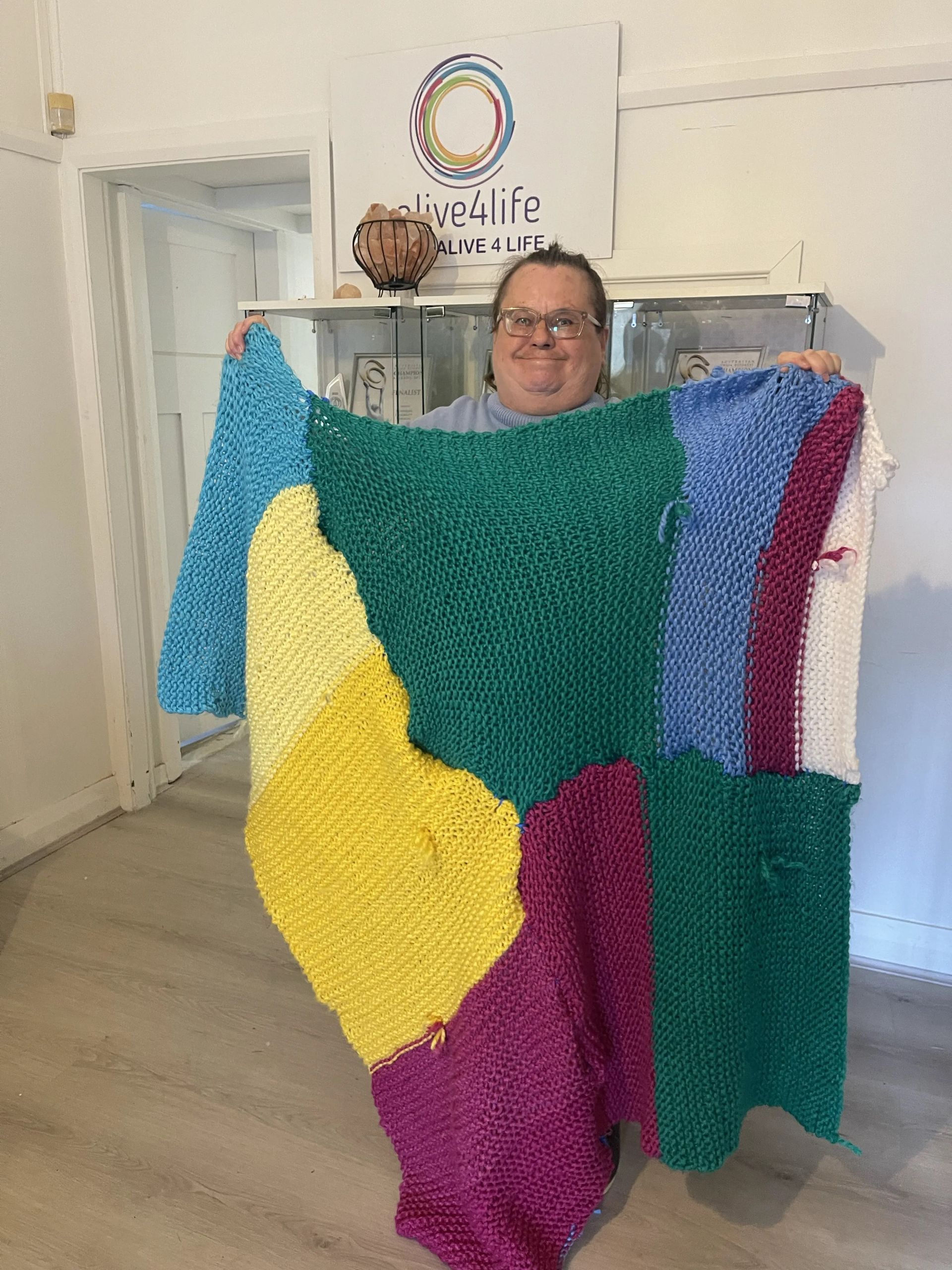 Alive4Life participant holding up a large multicoloured knitted blanket in front of the Alive4Life sign