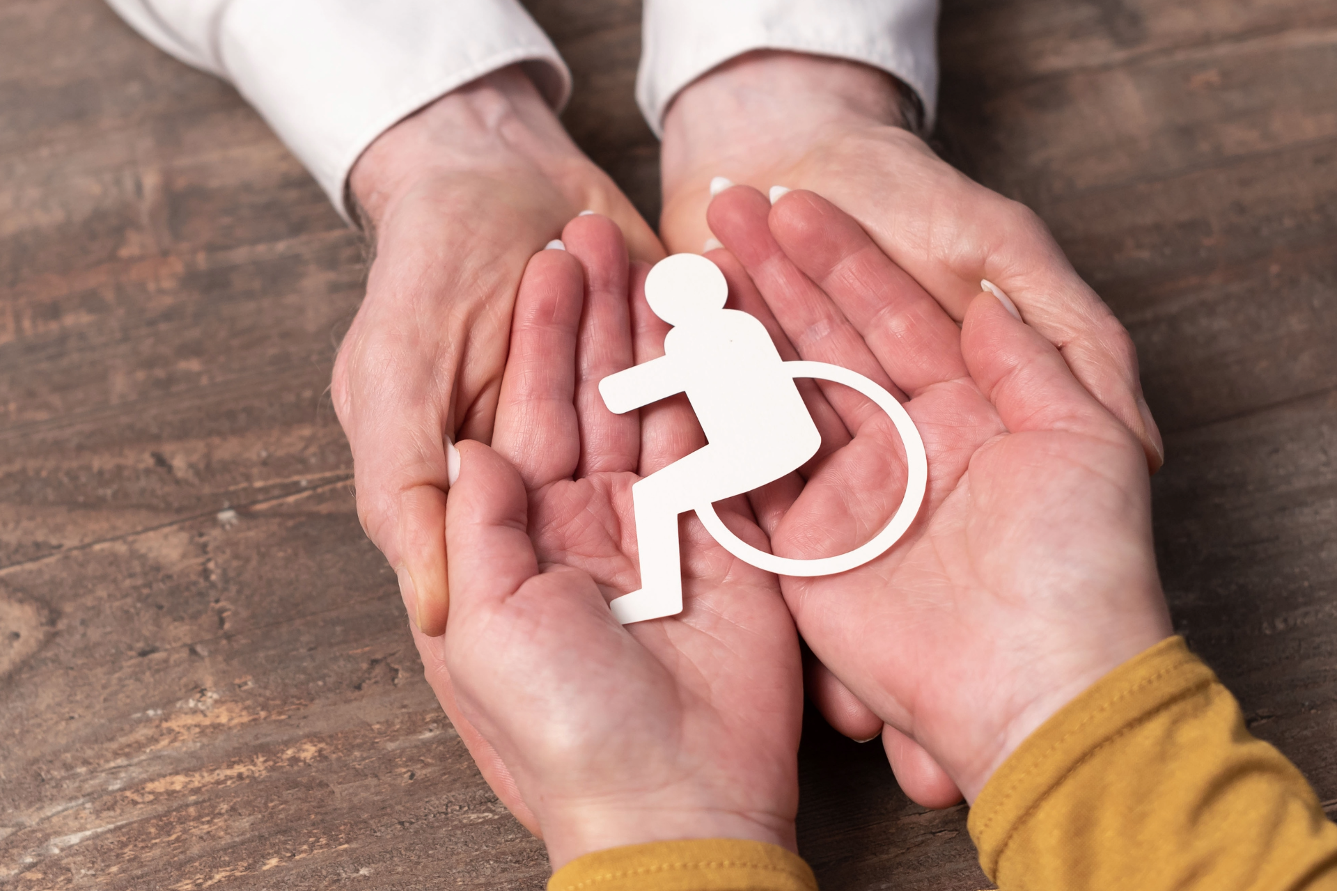 Hands holding a white wheelchair accessibility symbol on a wooden table.