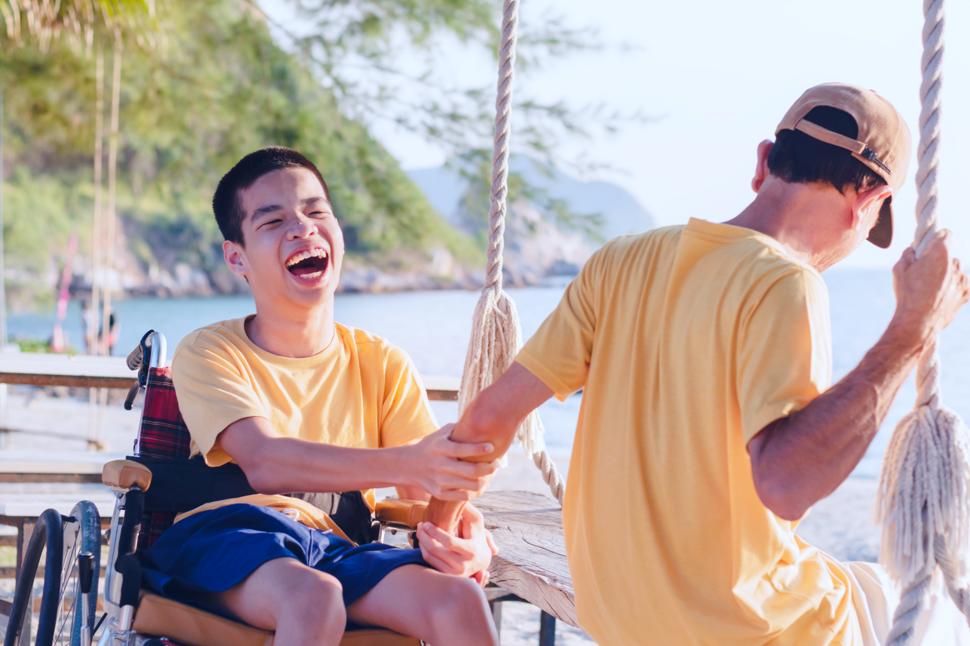 Two friends in yellow shirts laughing on a boat by the water, with a tropical shoreline in the background