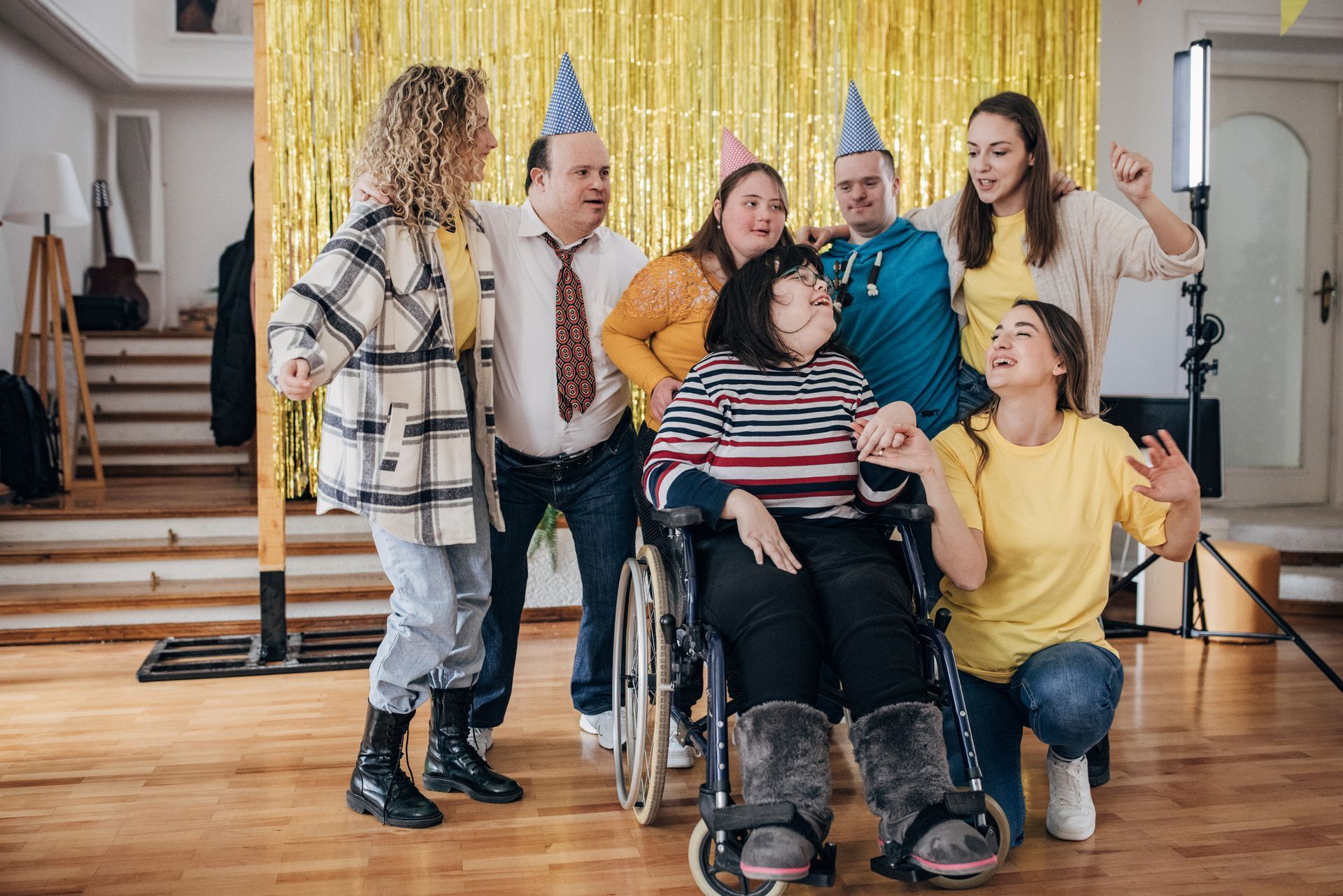 Group of people in party hats, one in a wheelchair, smiling in front of gold fringe backdrop.