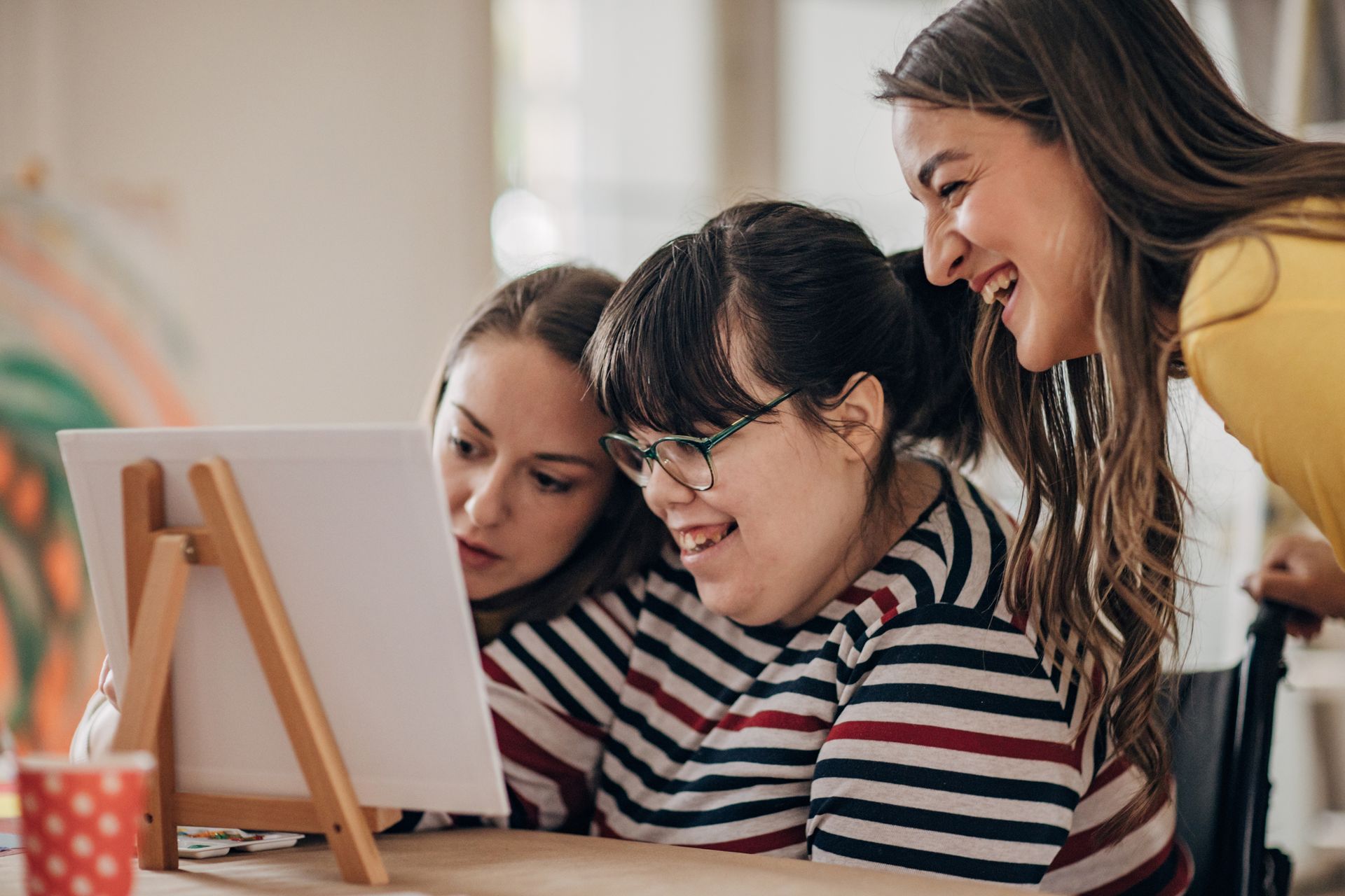 Three people looking at a painting on an easel. Two smile. Indoors, art studio setting.