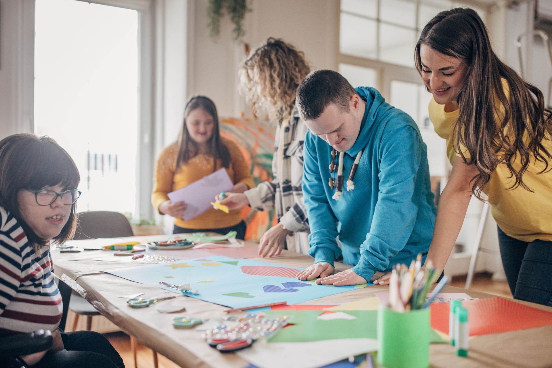 People with disabilities creating art at a table with assistance, indoors.
