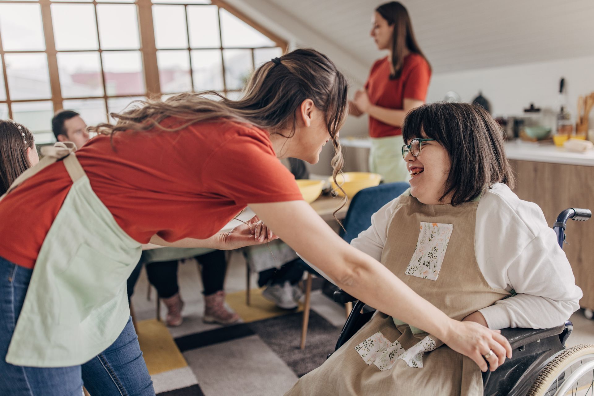 Woman in red shirt assisting person in wheelchair with apron in a kitchen, smiling.