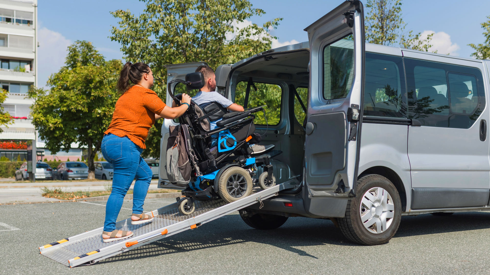 Two people help a person in a wheelchair up a ramp into a gray van outdoors.