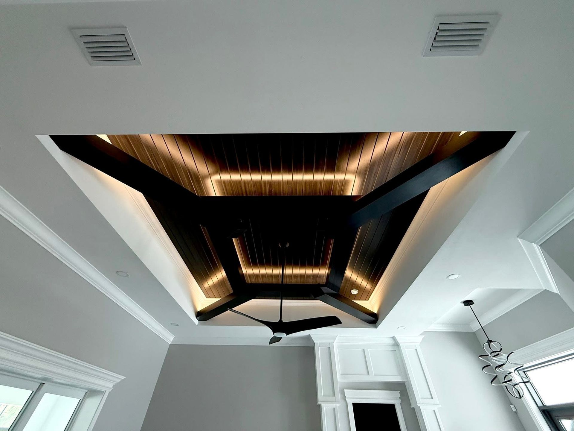 A modern tray ceiling with dark wood slats, LED accent lighting, and a black ceiling fan in a white room.