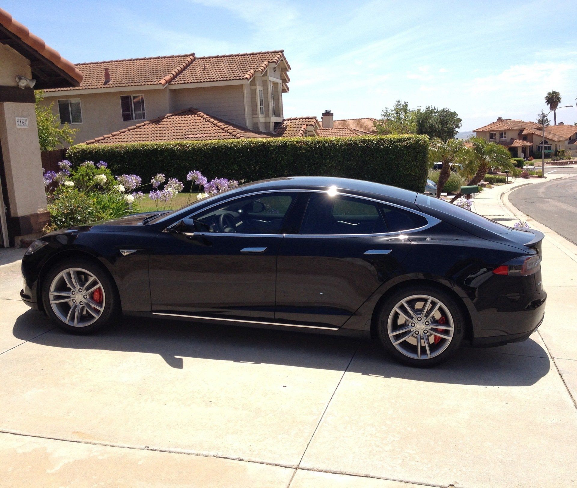 Black Tesla Model S parked on a driveway in front of a house on a sunny day.