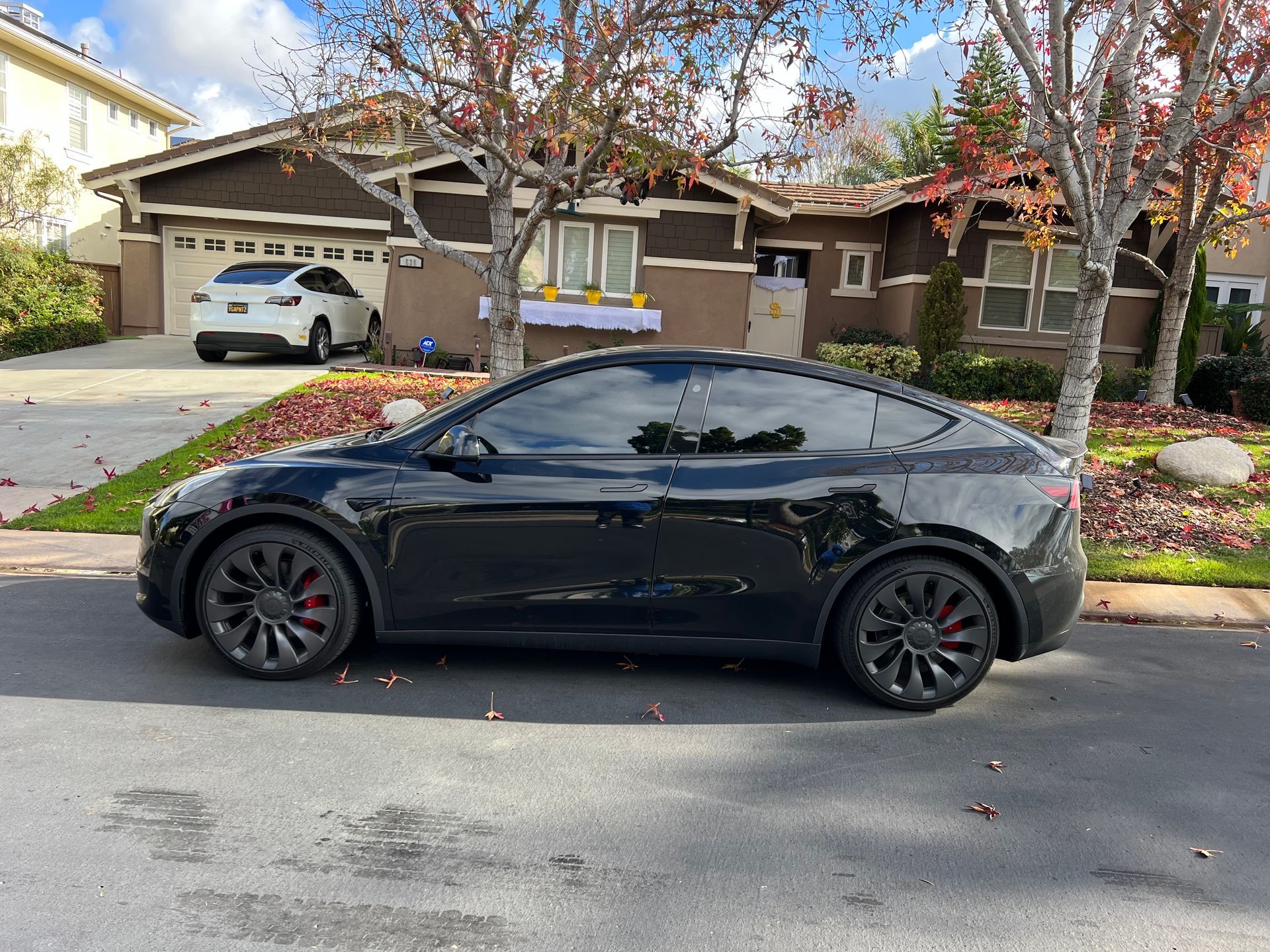 Black Tesla Model Y parked on a street in front of a house, another Tesla parked in the driveway.