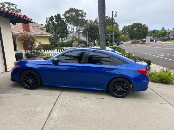 Blue sedan parked on a driveway with black rims, in front of a house on a cloudy day.
