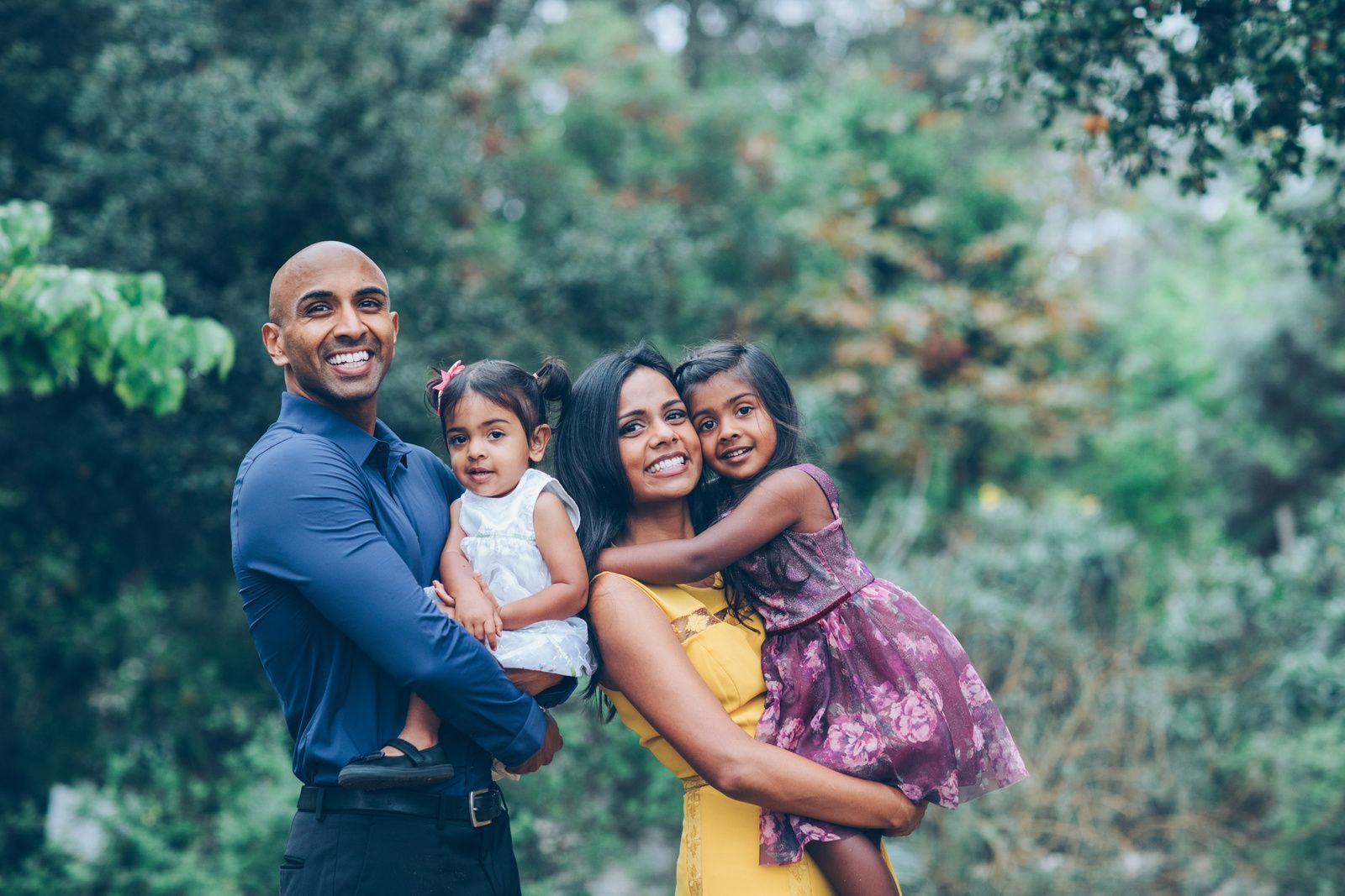 Smiling family of four outdoors, with two children being held in a leafy park.