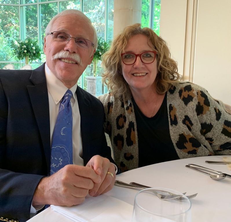 Two smiling people seated at a restaurant table, with a window and greenery behind them.