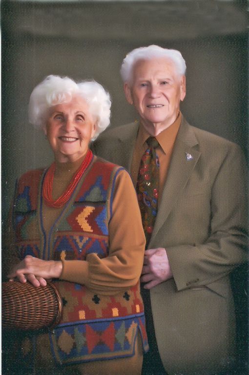 Two people posing indoors, one in a patterned sweater and the other in a suit, both smiling.