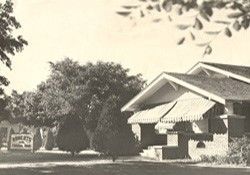 Black-and-white photo of a house with a striped awning, trees, and a roadside sign.