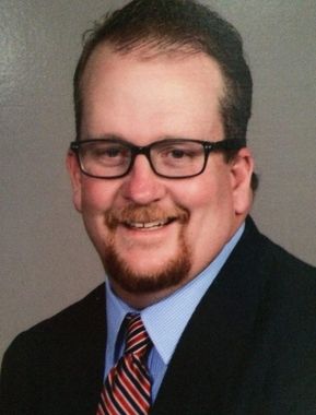 Smiling man in glasses, dark suit, blue shirt, and striped tie against a gray background