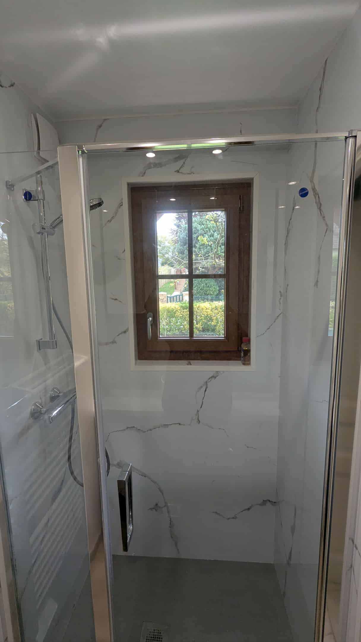 Cabine de douche étroite carrelée avec des murs imitation marbre et une petite fenêtre au fond.