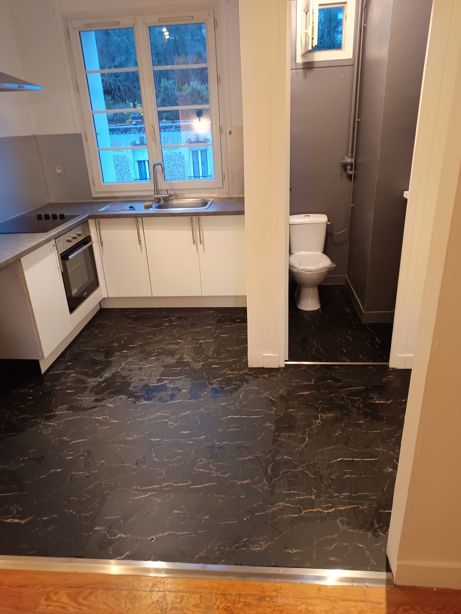 Petite cuisine avec placards blancs, sol en marbre noir et toilettes au fond.