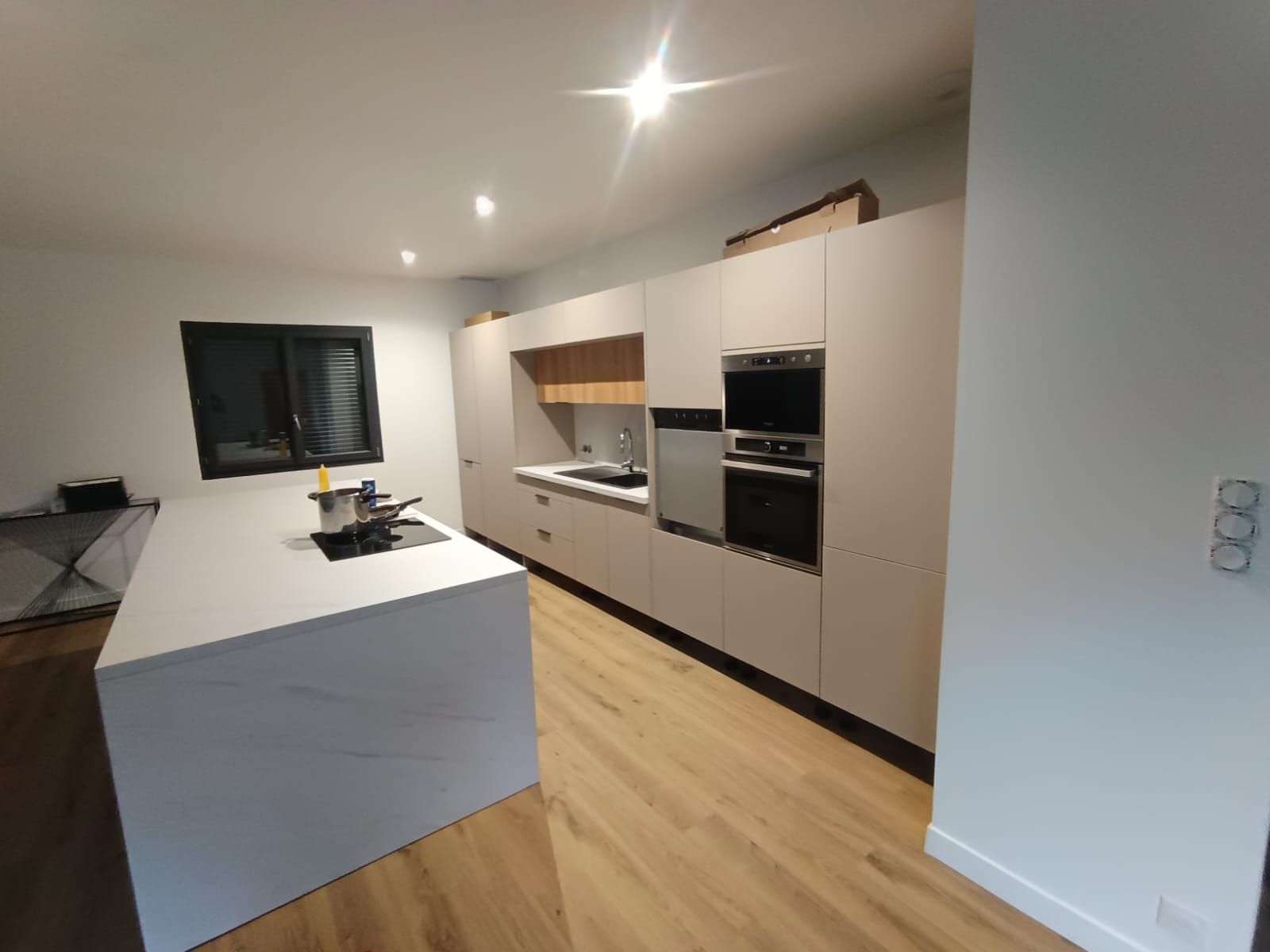 Cuisine moderne blanche avec îlot central, fours encastrés, parquet et stores noirs.
