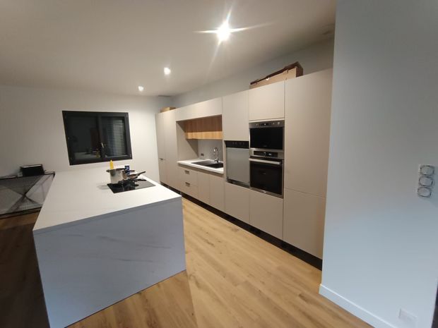 Cuisine moderne avec îlot central blanc, parquet, four encastré et grande fenêtre à cadre noir.