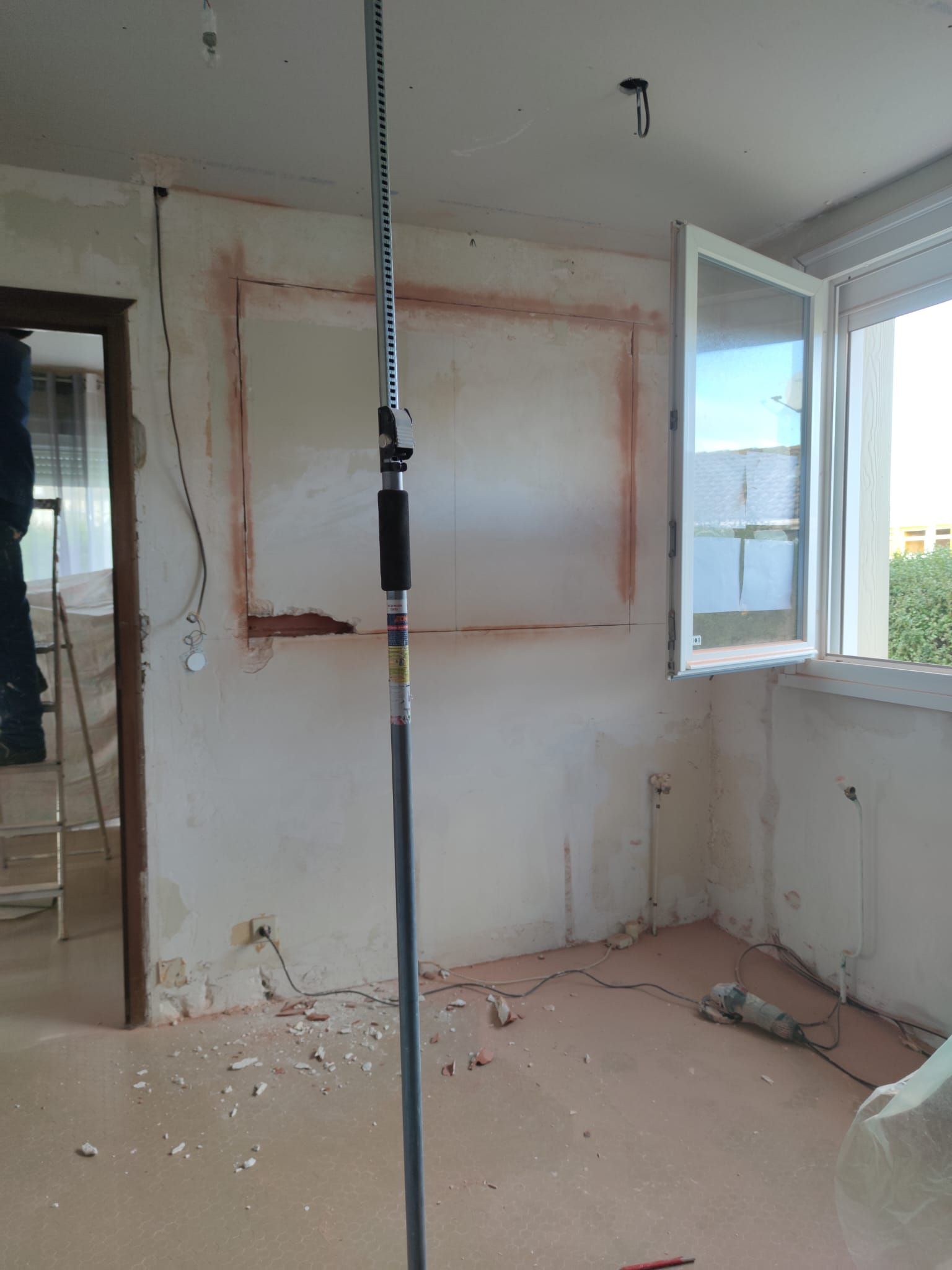 Chambre partiellement rénovée avec fenêtre ouverte, murs nus et débris de construction au sol