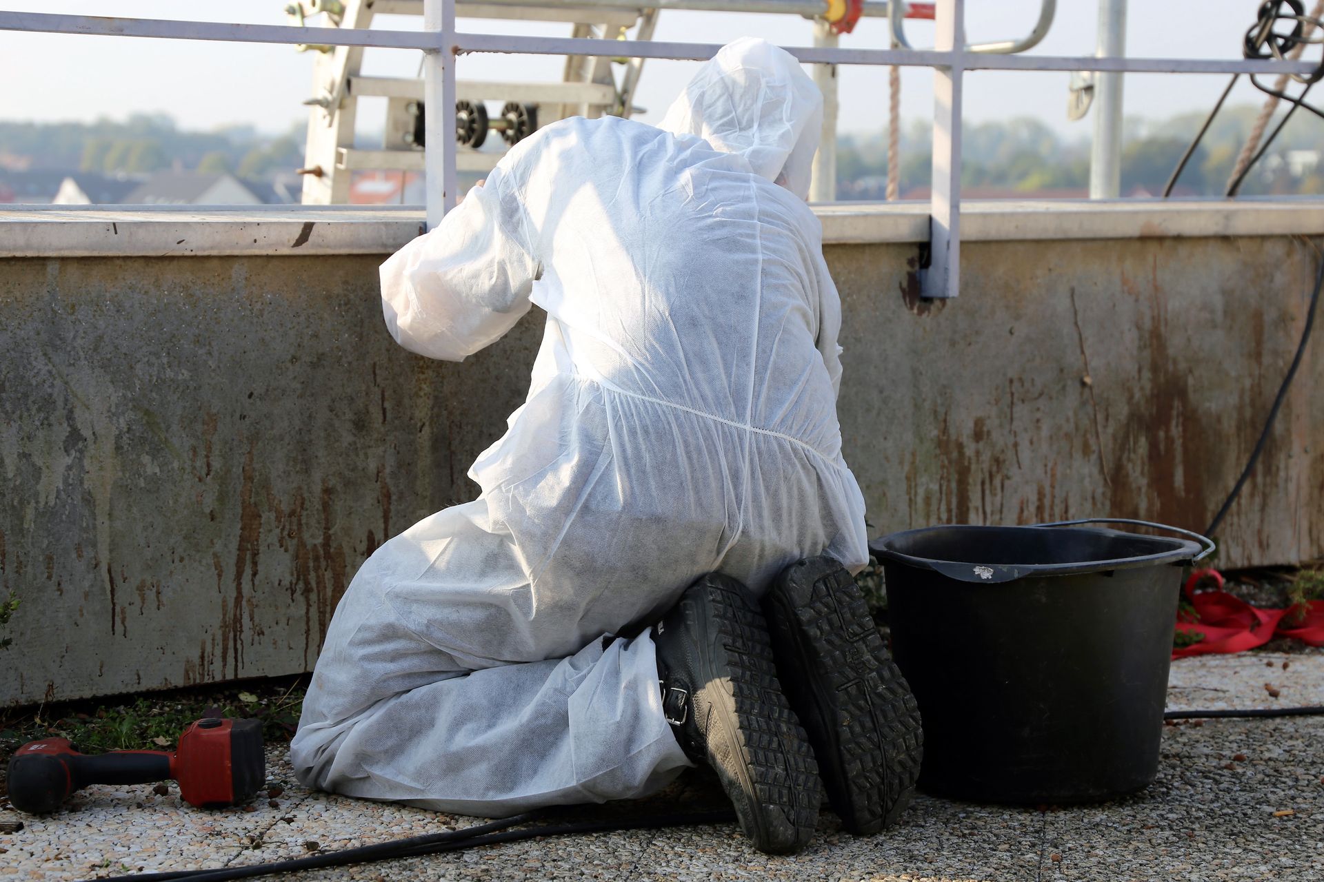 Person in white protective suit kneeling near a wall, working, with a bucket and tools nearby.