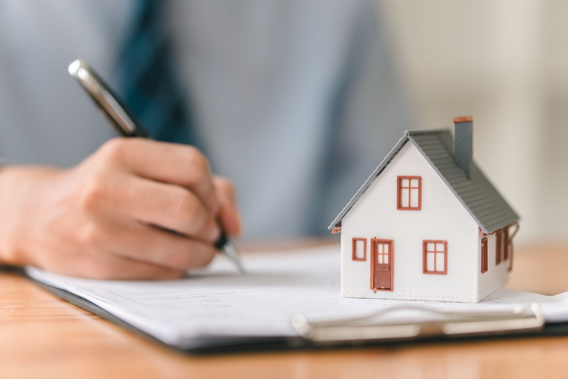 Hand writing on a clipboard beside a small model house, suggesting a home inspection or real estate appraisal