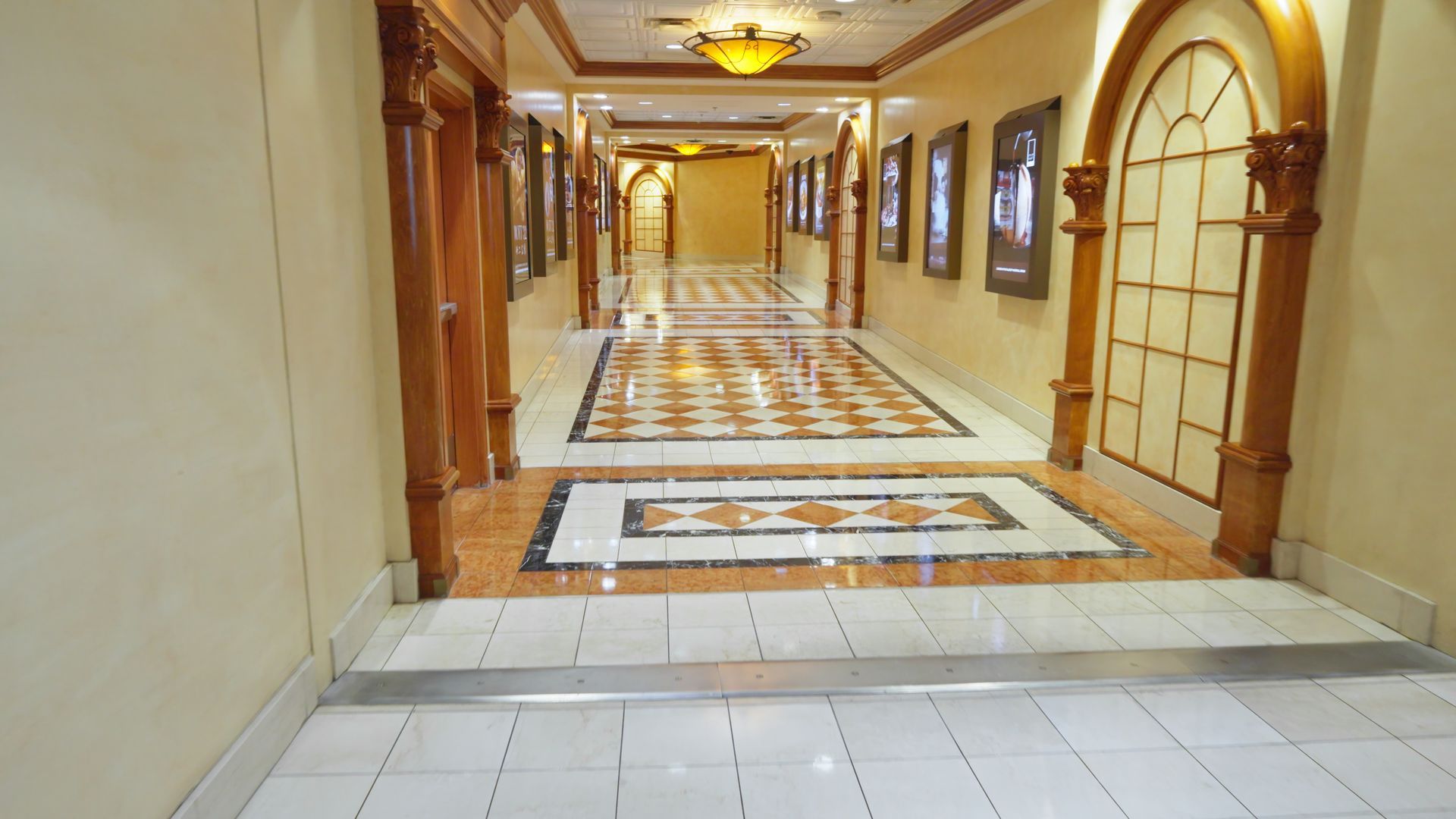 Empty hotel hallway with beige walls, arched doorways, patterned carpet, and framed wall art