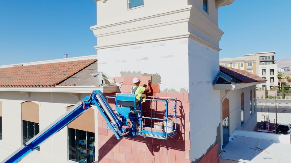 Worker in a blue lift paints the exterior wall of a white building under a clear sky.