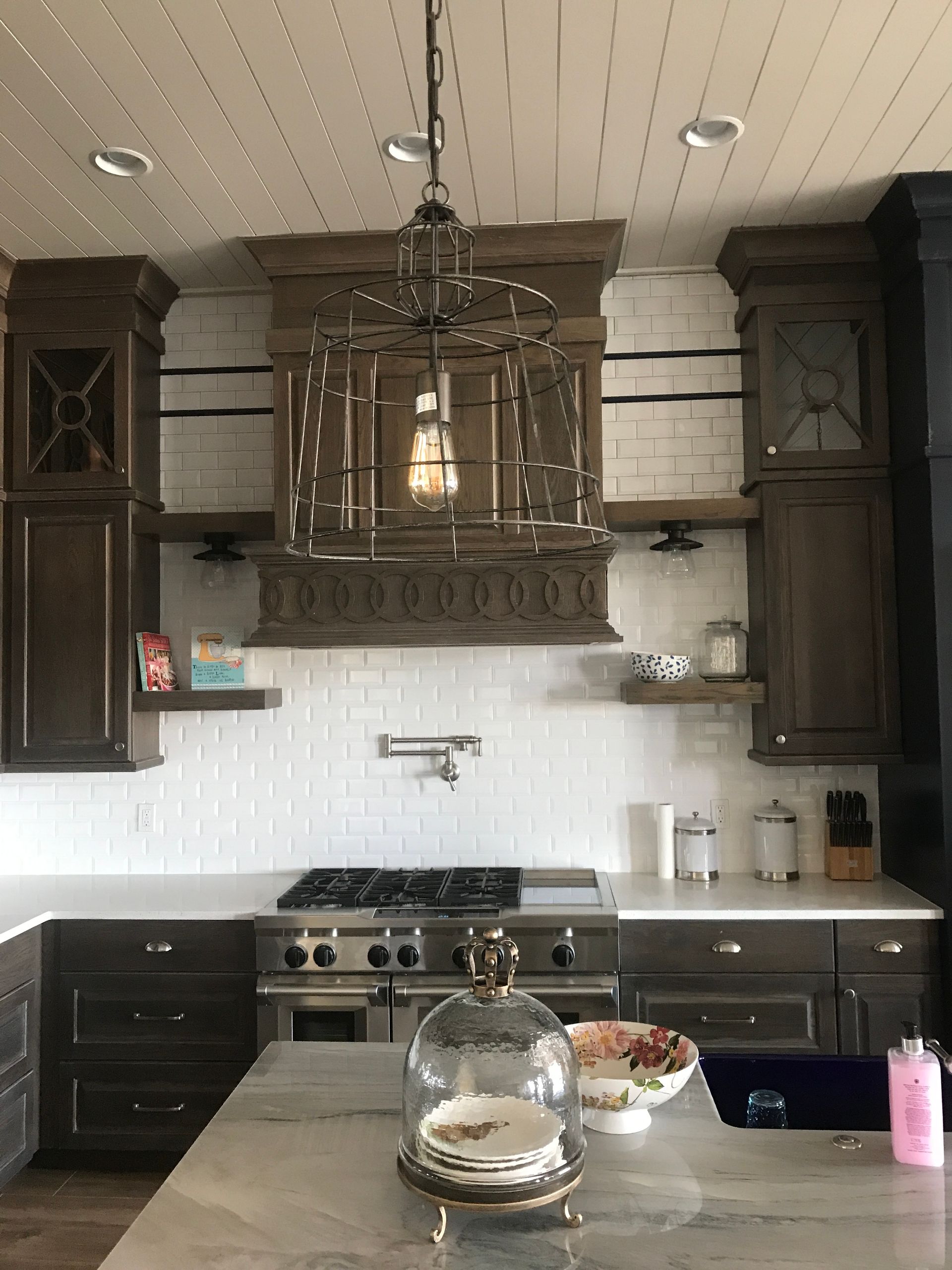 Kitchen with dark wood cabinets, stainless stove, large ornate hood, and island with a glass cloche and fruit bowl.