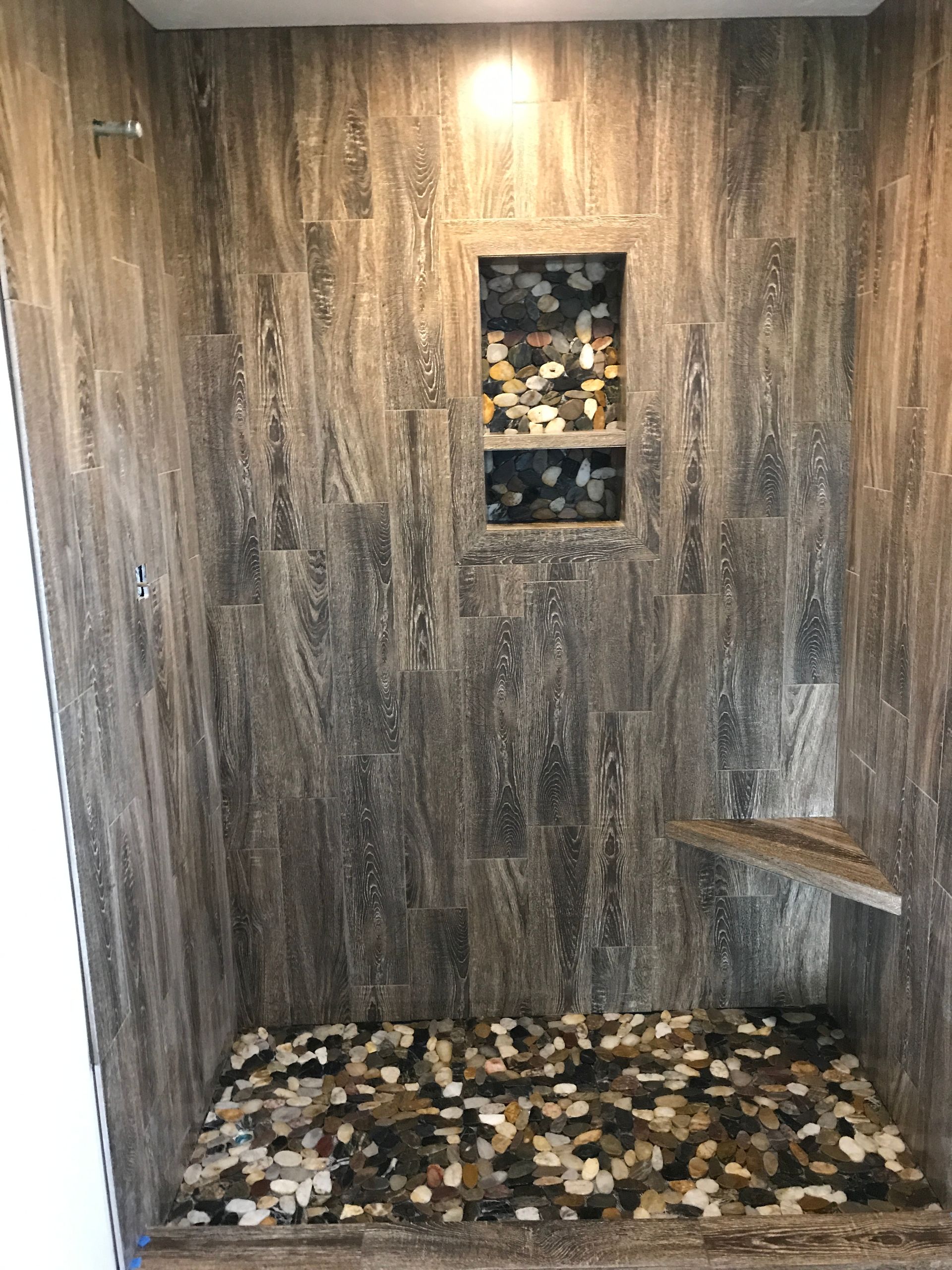 Empty tiled shower with wood-look walls, pebble floor, and a small built-in shelf niche.
