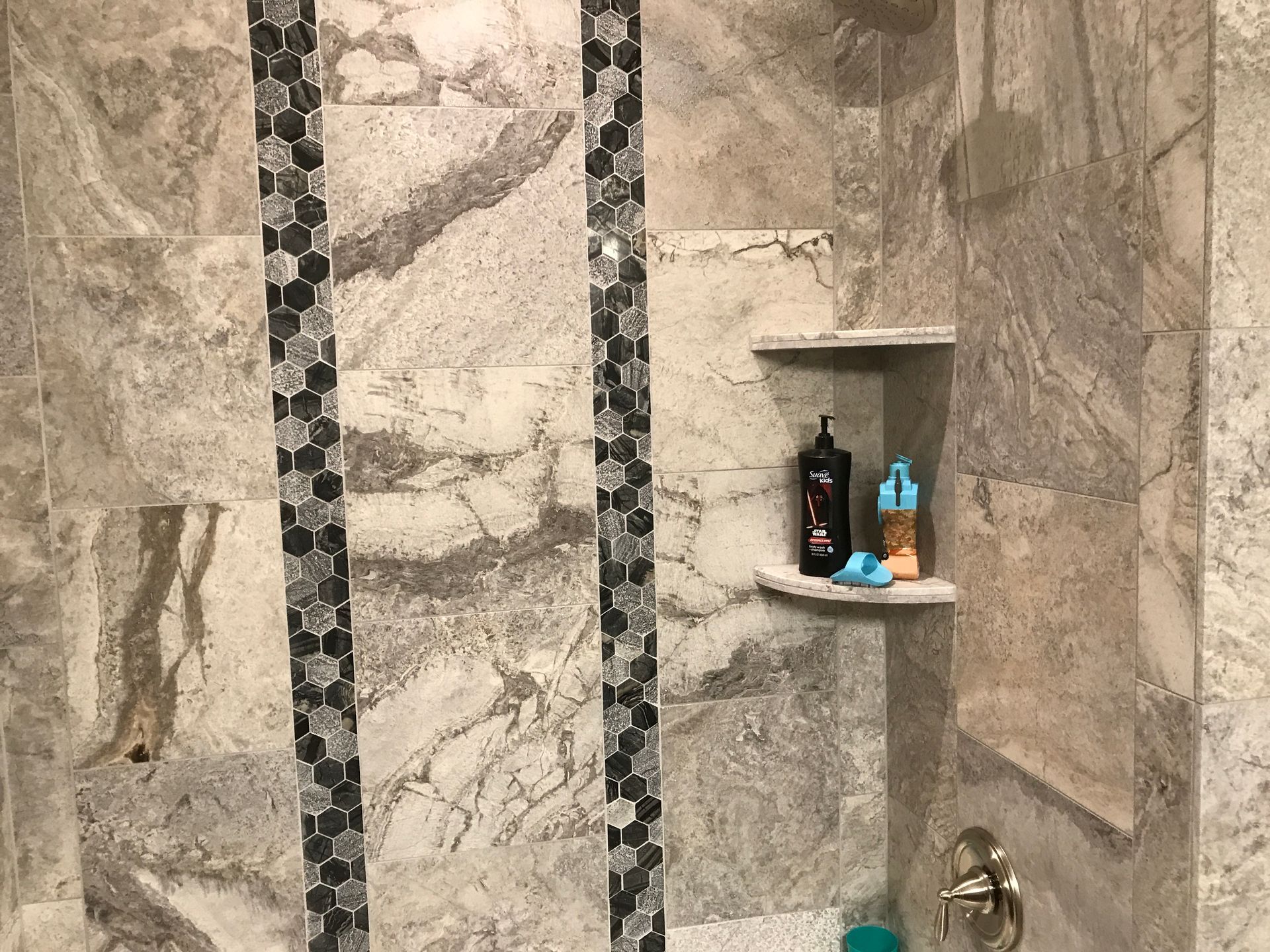 Shower with gray marble tile, built-in shelf holding bottles, and a black mosaic accent strip