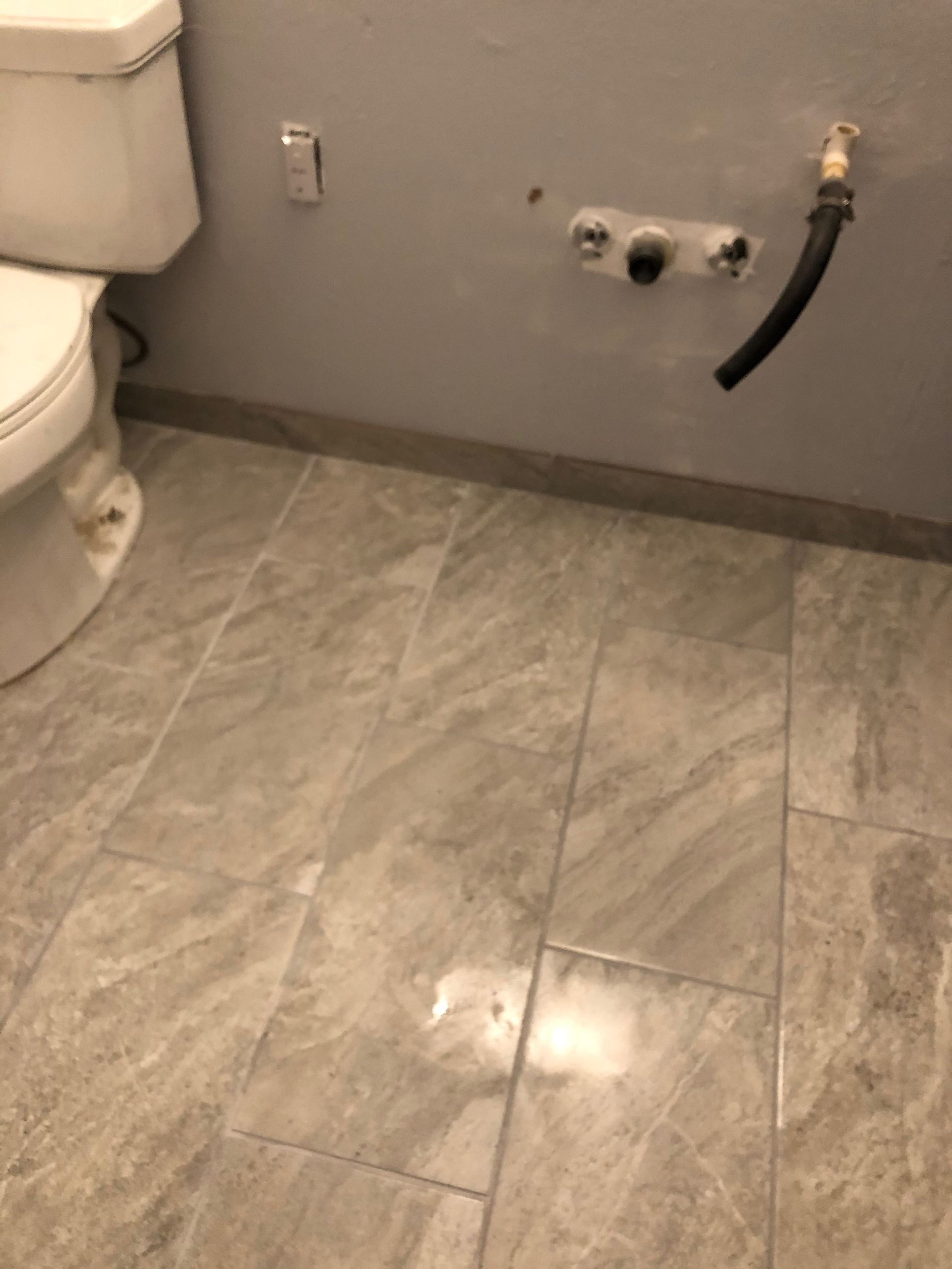 Bathroom floor with toilet, gray tile wall, and exposed plumbing hookup on the right.