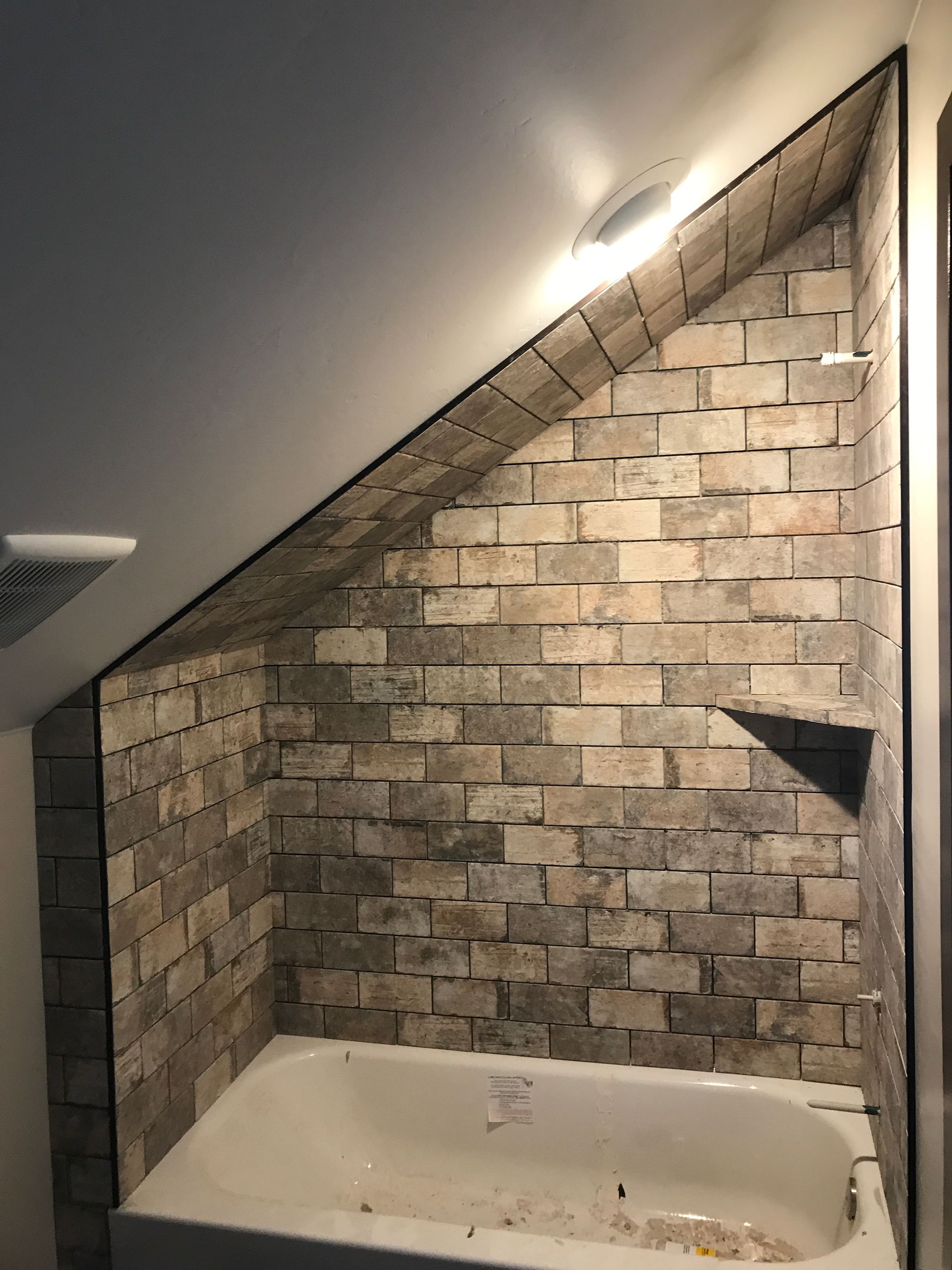 Brick-lined shower nook with a slanted ceiling and bathtub under a wall light
