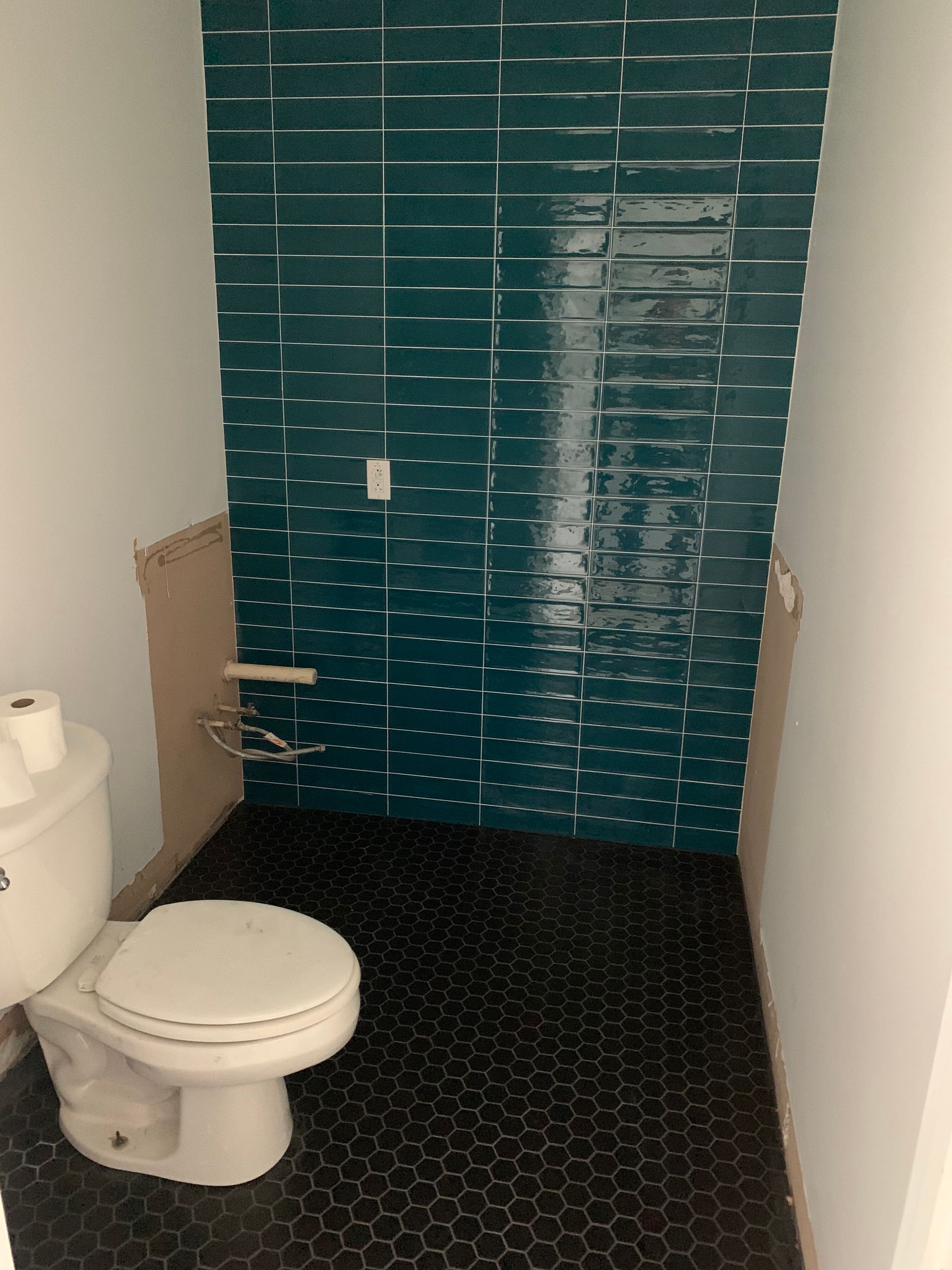 Small bathroom with toilet, black hex tile floor, and teal tiled shower wall with shelves.