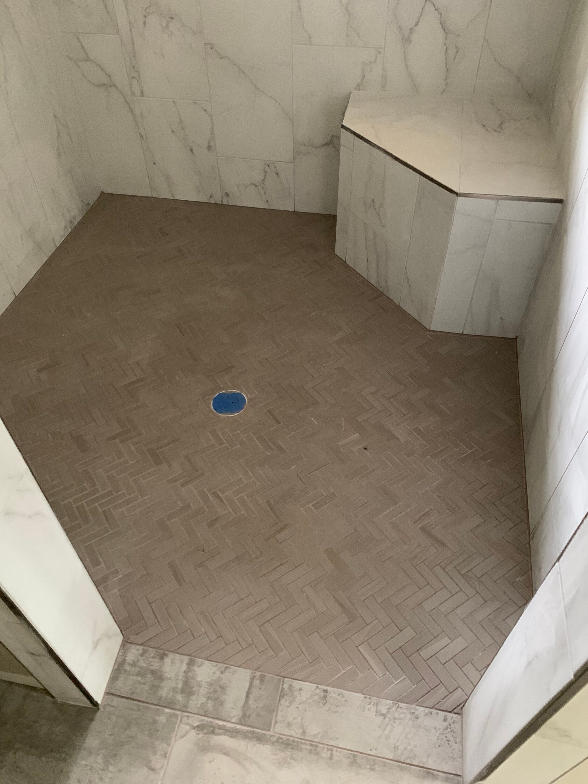 Small empty tiled shower stall with beige floor, marble-look walls, and a blue floor drain.