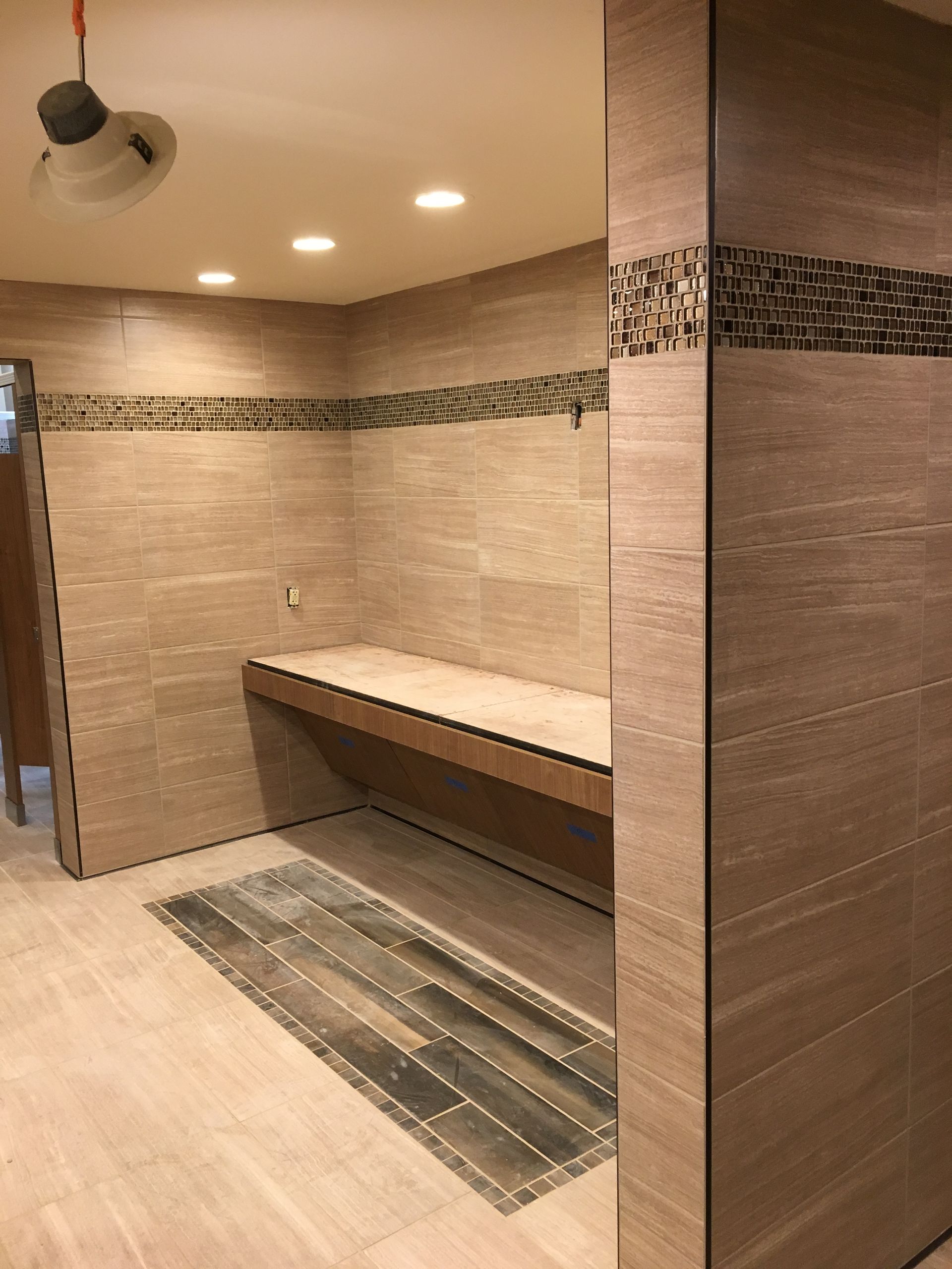 Empty tiled sauna changing area with a wooden bench and floor mat under recessed lights