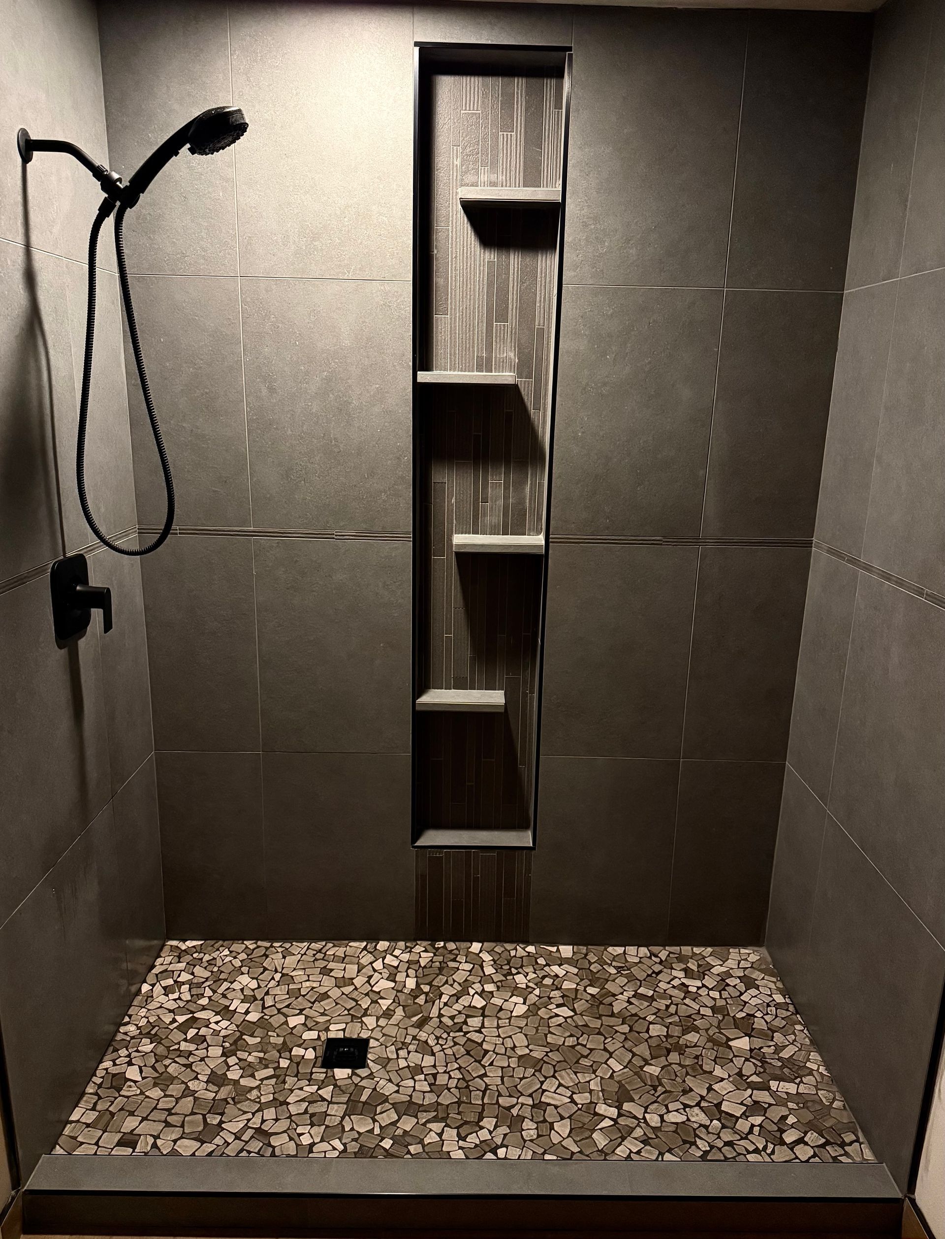 Small tiled shower with a wall niche, handheld showerhead, and pebble-patterned floor.