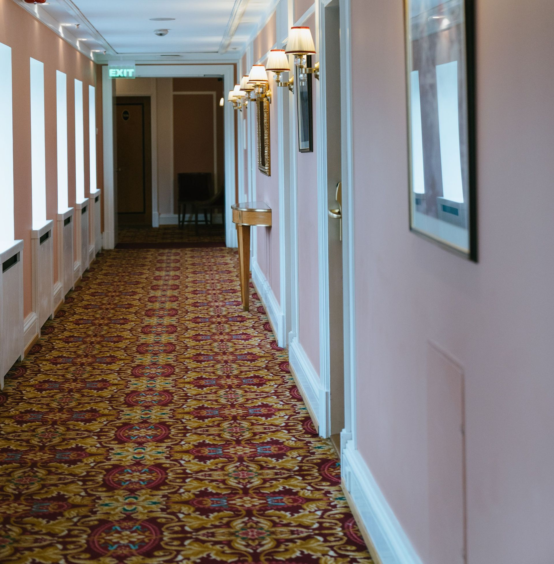Carpeted hotel hallway with pink walls, framed art, and wall sconces