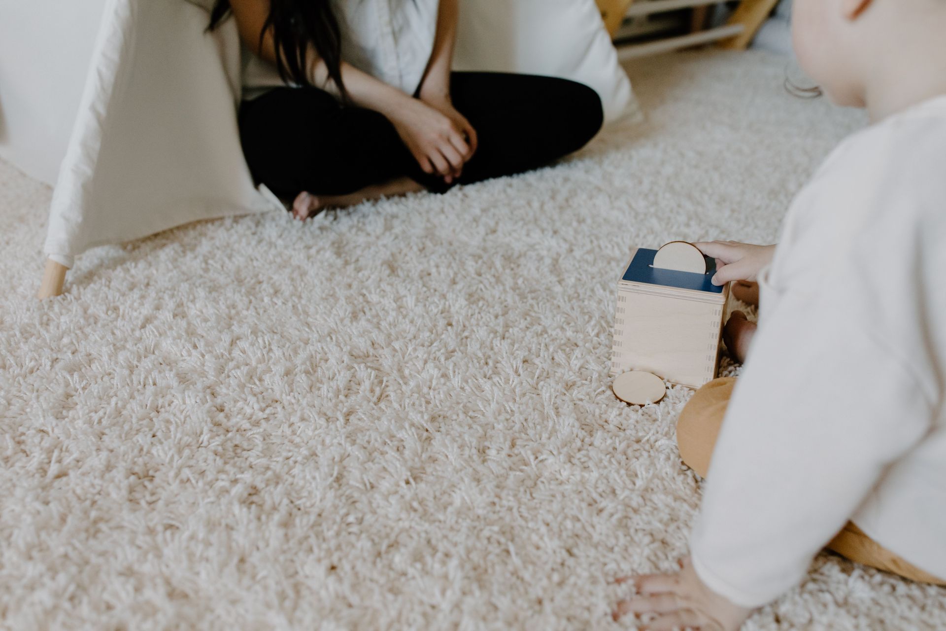 Two people sitting on a fluffy white rug, playing with a small wooden toy house